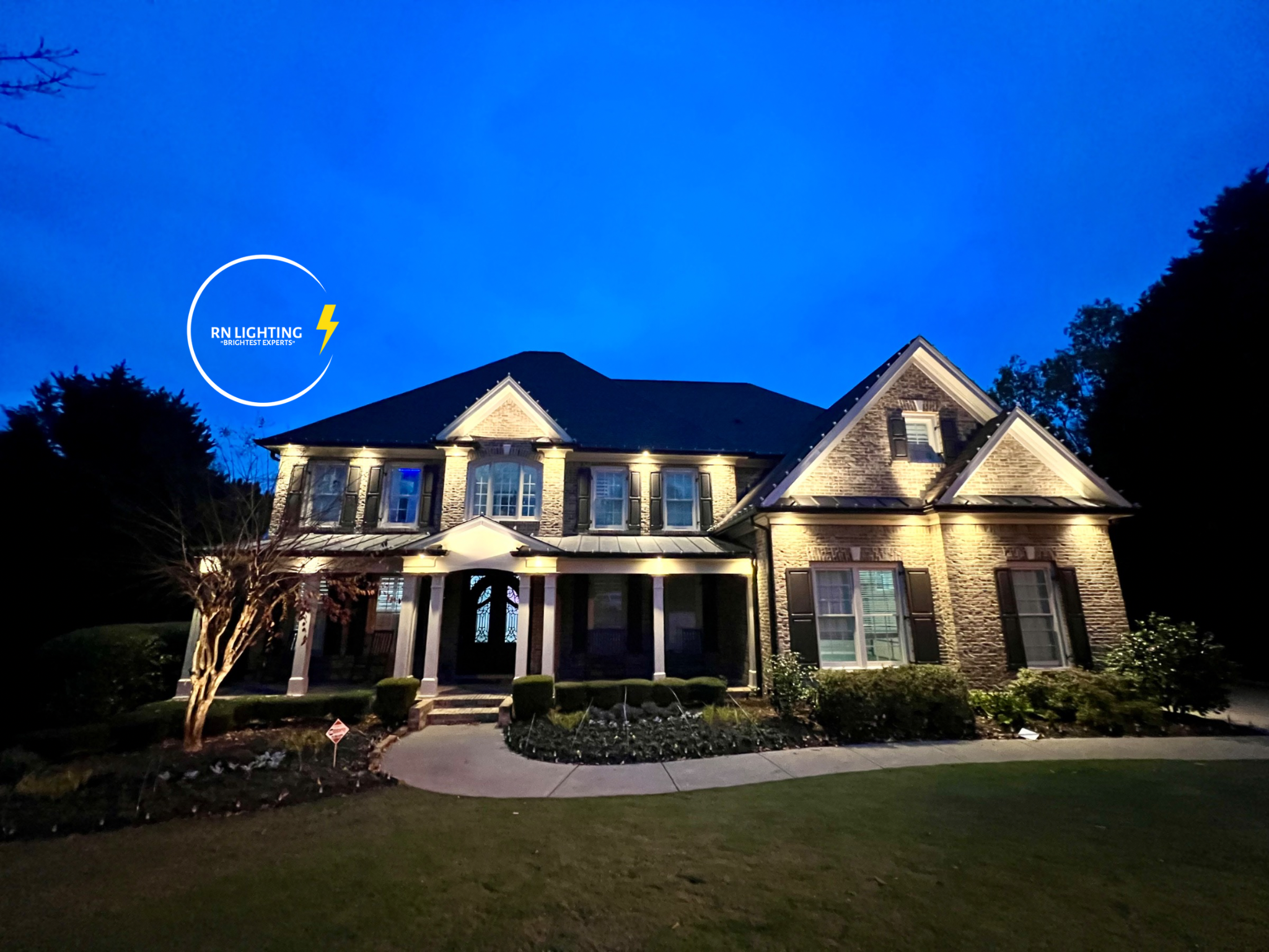 A two-story brick house illuminated at night with outdoor lights, a front lawn, a curved pathway, and a small garden with shrubs and trees.