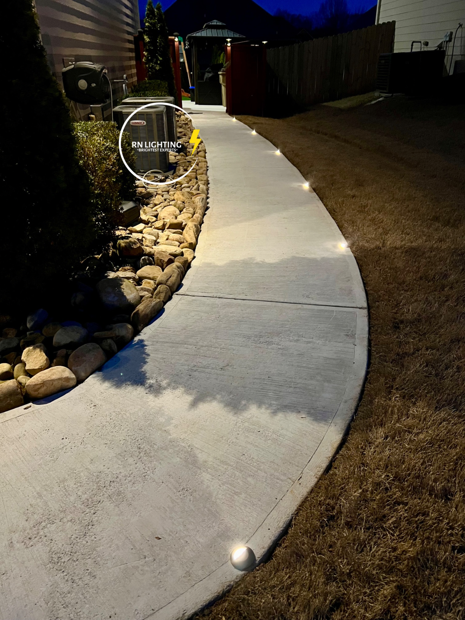 Nighttime outdoor scene with a concrete sidewalk, small ground lights along the side, stone landscaping with bushes, HVAC unit marked 'RN LIGHTING', and a fenced yard with a shed.