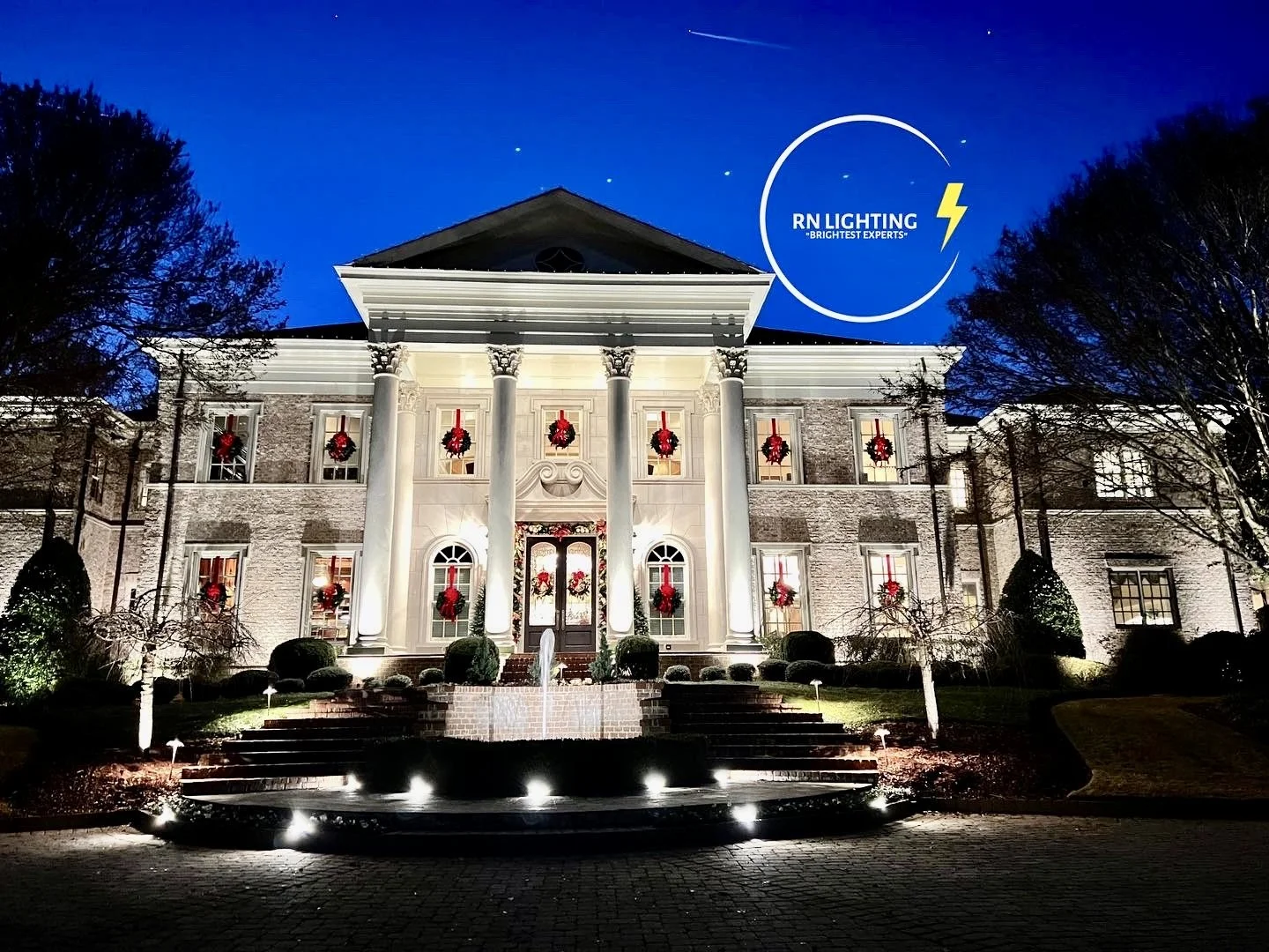 A large, elegant, white house decorated with Christmas wreaths and red ribbons, illuminated at night with landscaping and a small fountain in front, with a blue sky overhead featuring a logo for RN Lighting.