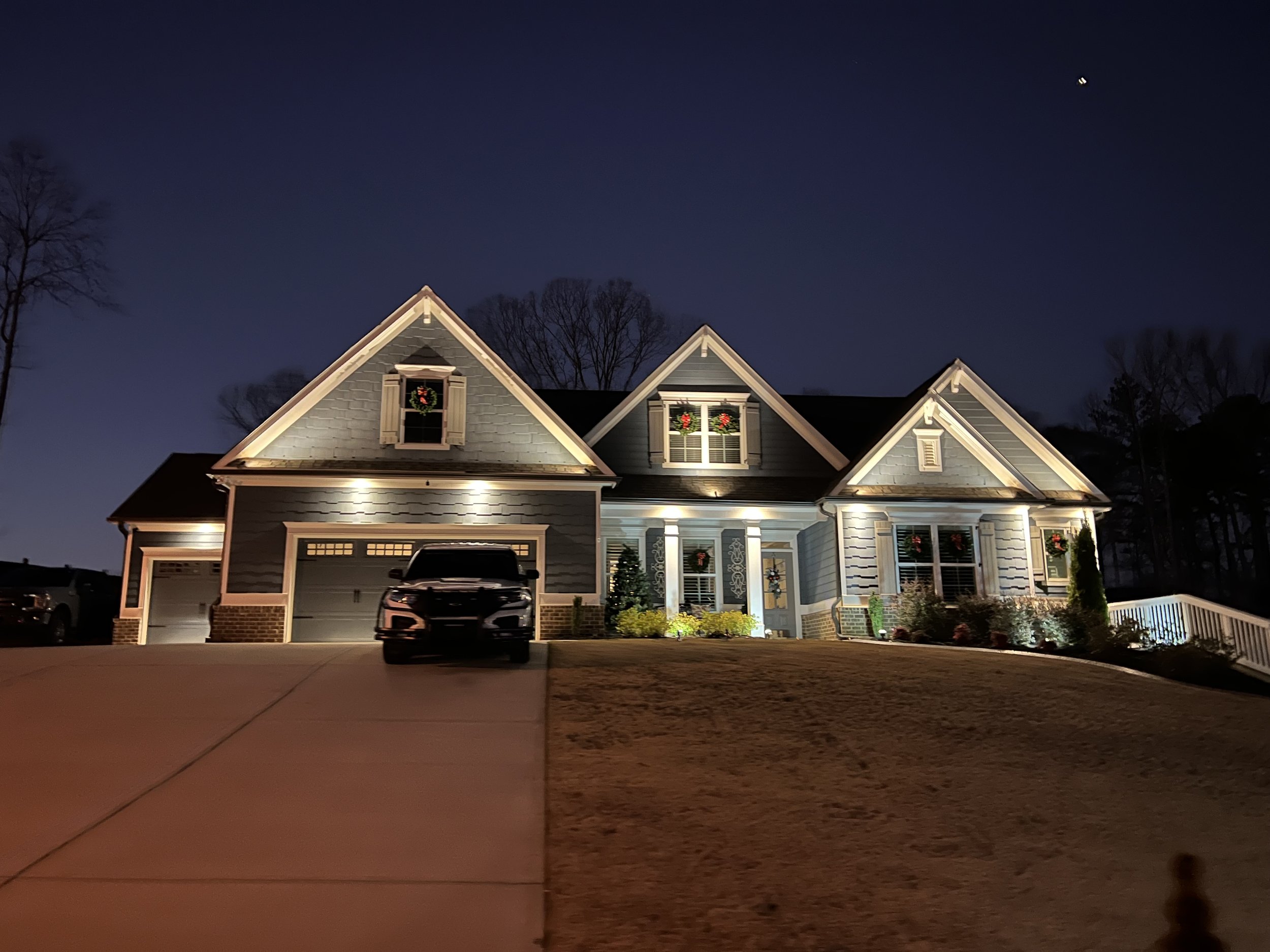 A house decorated with Christmas wreaths in the windows at night, with outdoor lighting illuminating the facade and a vehicle parked in the driveway.