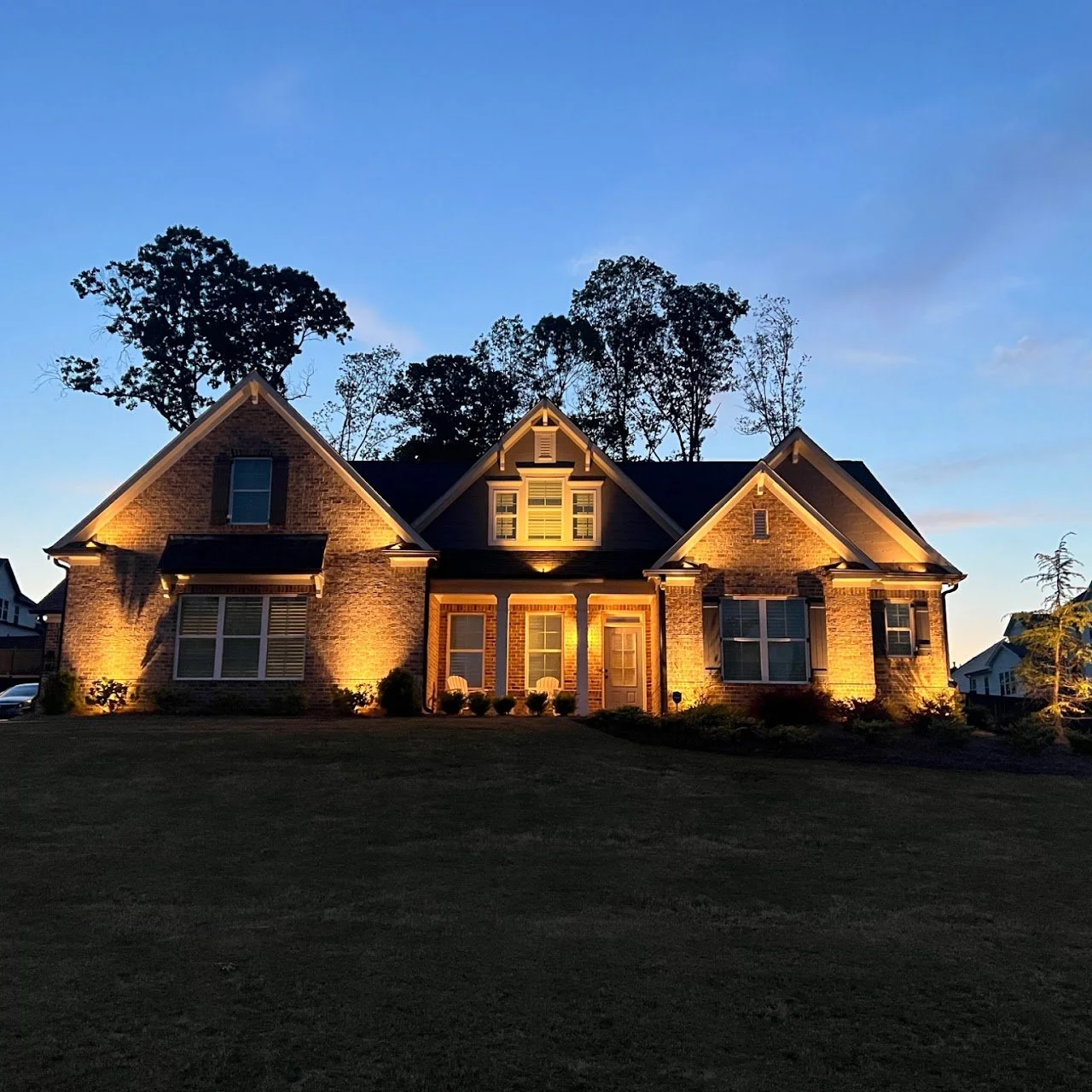 A large brick house illuminated at dusk with outdoor lighting, surrounded by a grassy lawn and trees in the background.