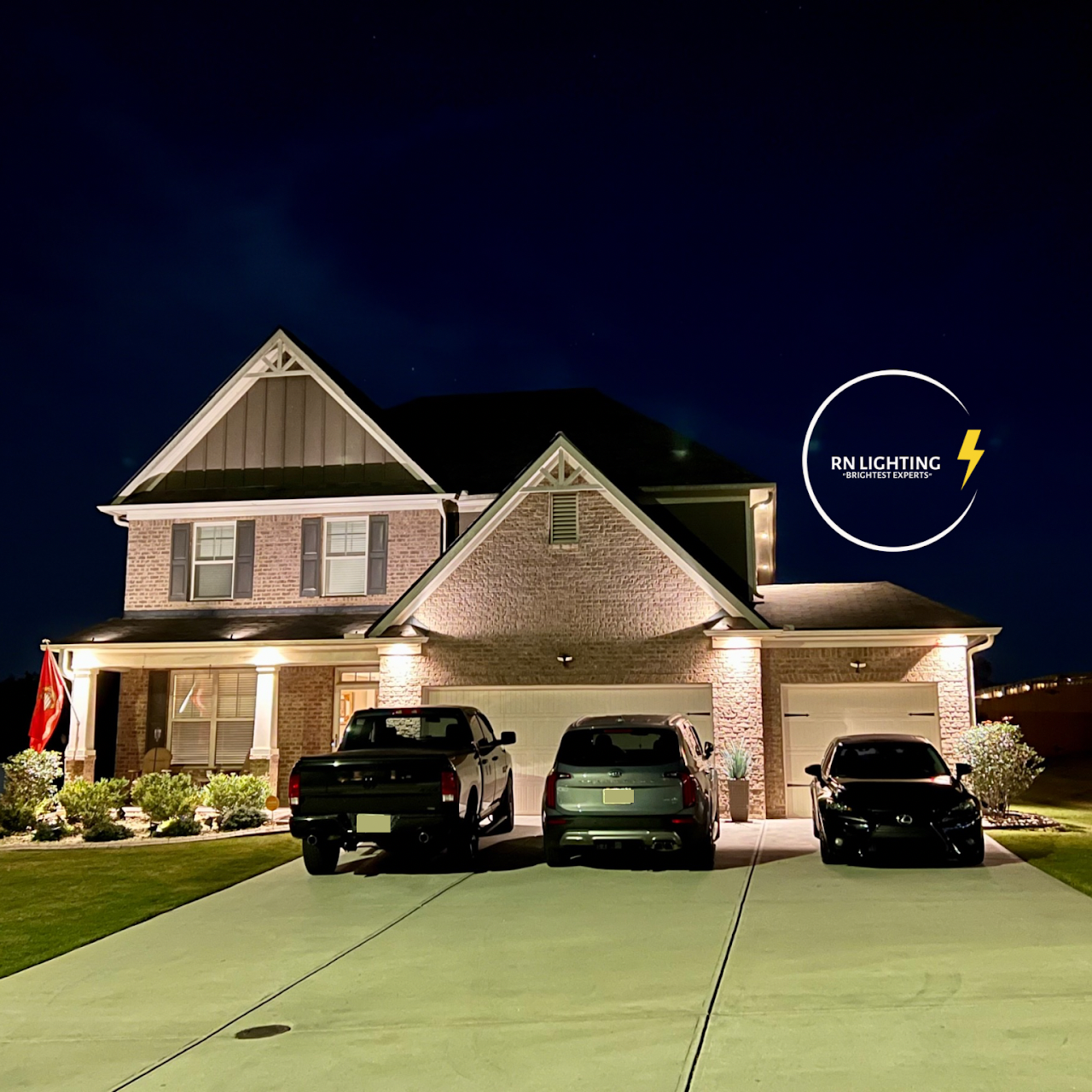 A two-story brick house at night with exterior lights on, three parked cars in the driveway, landscaped bushes, a flag on a pole, and a logo for RN Lighting in the sky.