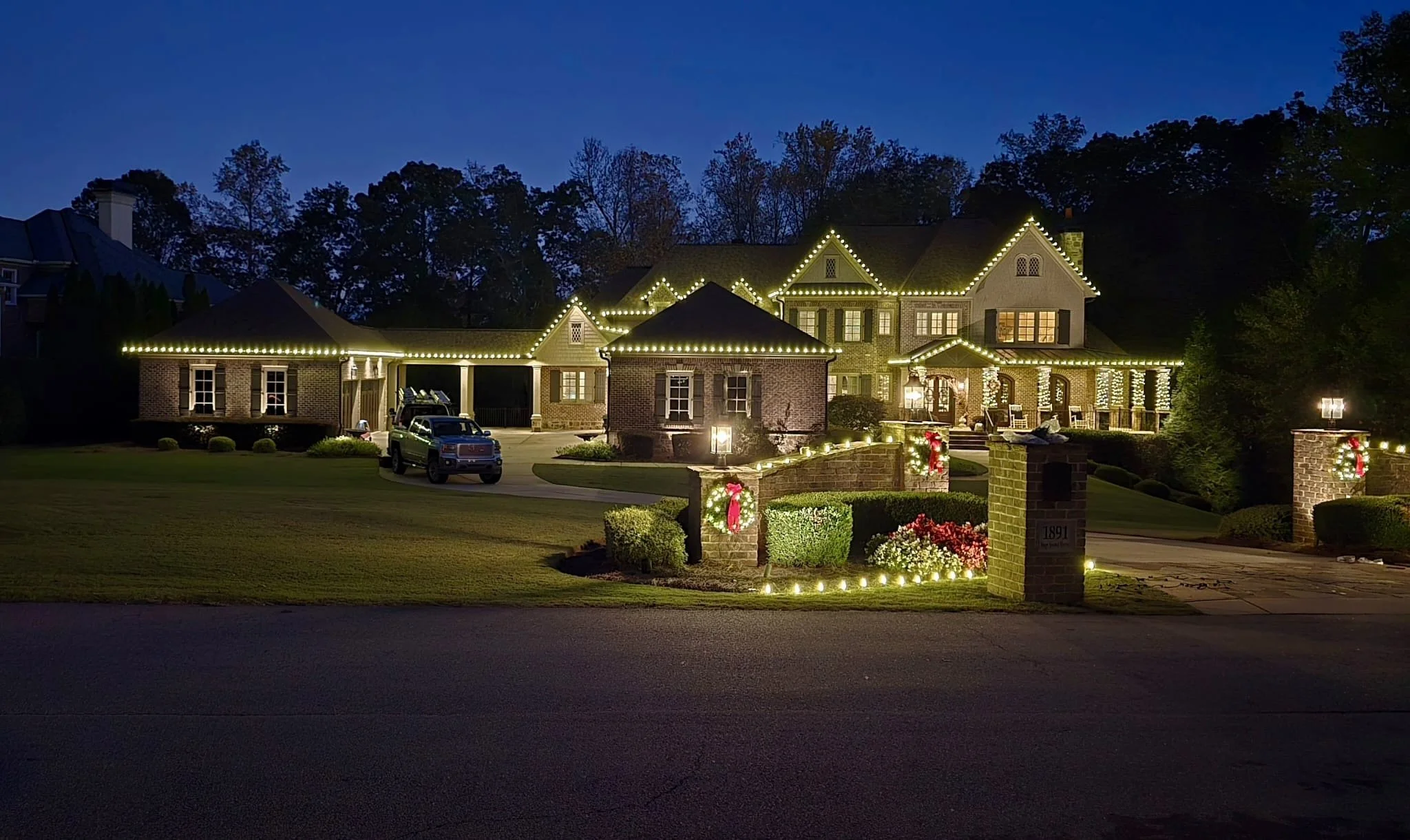 A large house decorated with string lights for Christmas, with a well-kept lawn, a driveway with a pickup truck, and a brick and stone fence with holiday wreaths and lighting.