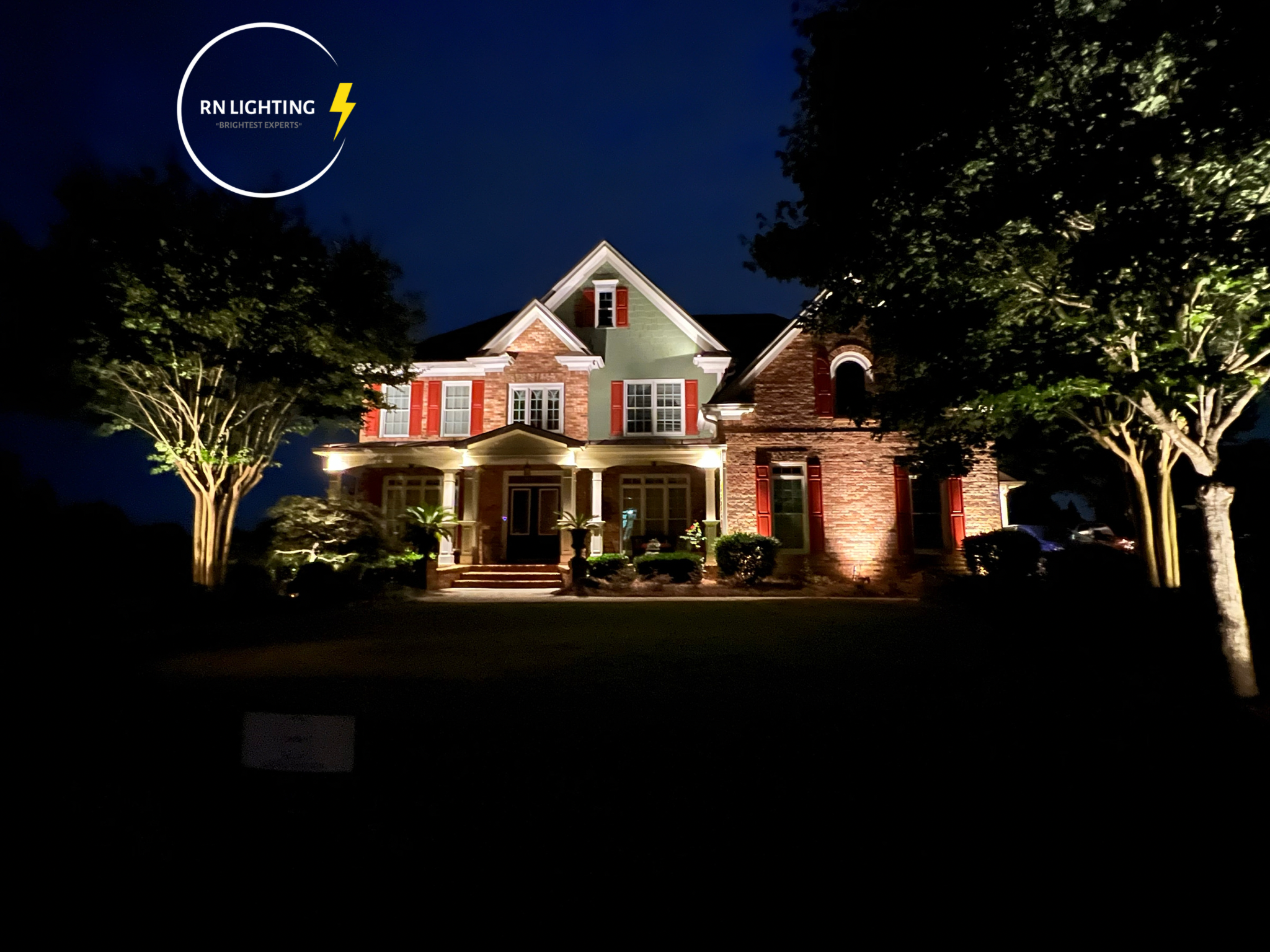 A large house illuminated at night, with trees on both sides and lights highlighting the front entrance.