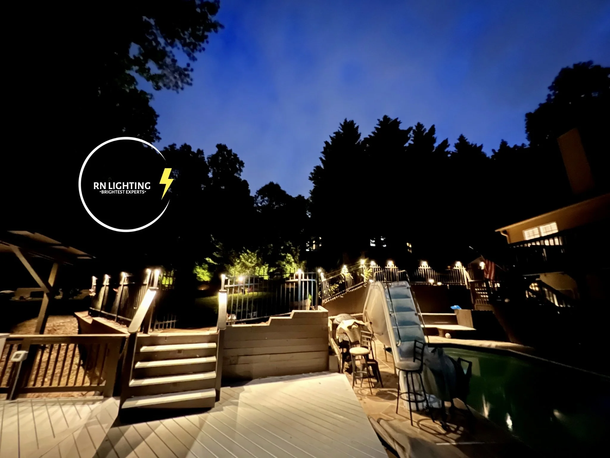 Nighttime view of a backyard patio with outdoor furniture, decking, trees, and a building, illuminated by outdoor lights. A logo in the bottom left corner reads 'RN Lighting, Brighter Experts,' with a yellow lightning bolt symbol.