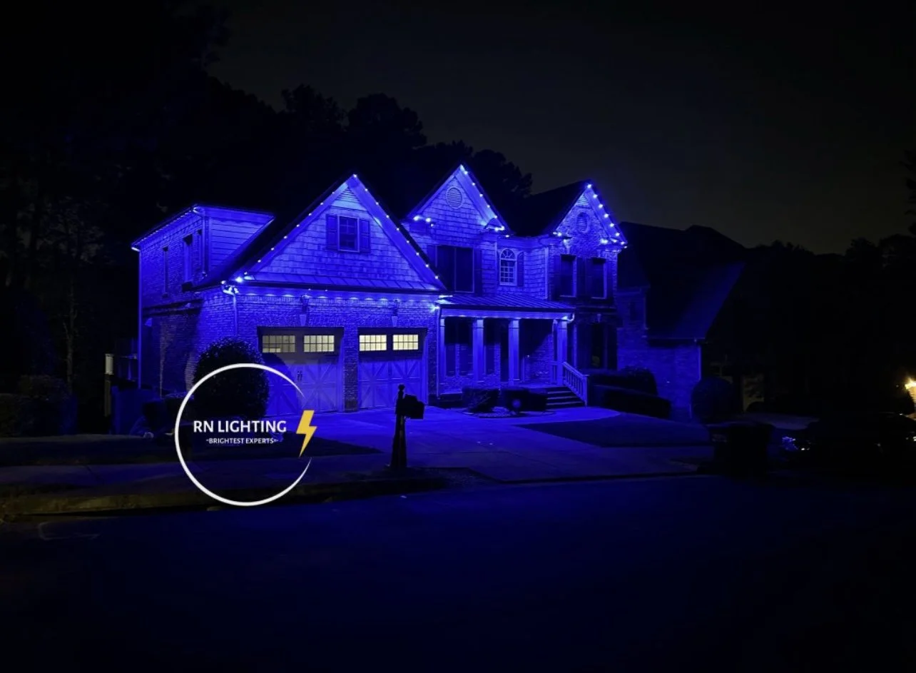 A large house illuminated with bright blue outdoor Christmas lights, with a sign reading 'RN Lighting Brightest Experts' in the front yard.