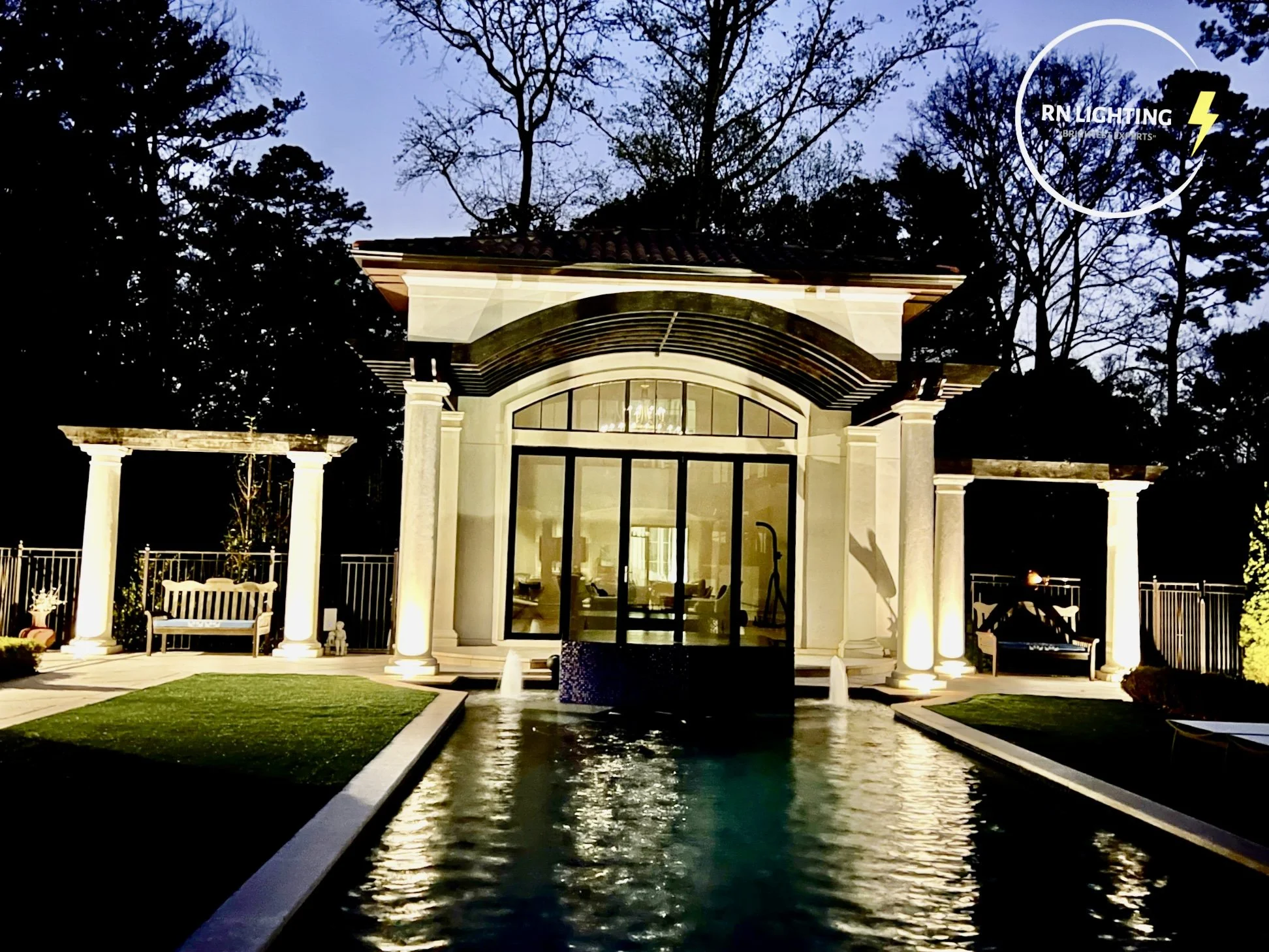 A backyard with a modern house, illuminated by outdoor lighting, featuring a water fountain, a pool, and decorative columns.