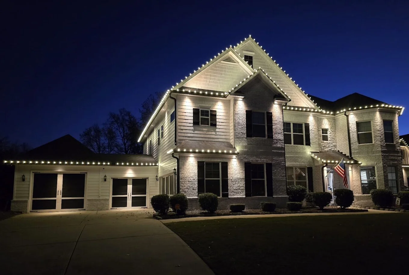 A large, two-story house decorated with string lights outlining its roof and windows at night. The house has white siding and stone accents, with an American flag near the front entrance and a driveway leading to a garage.