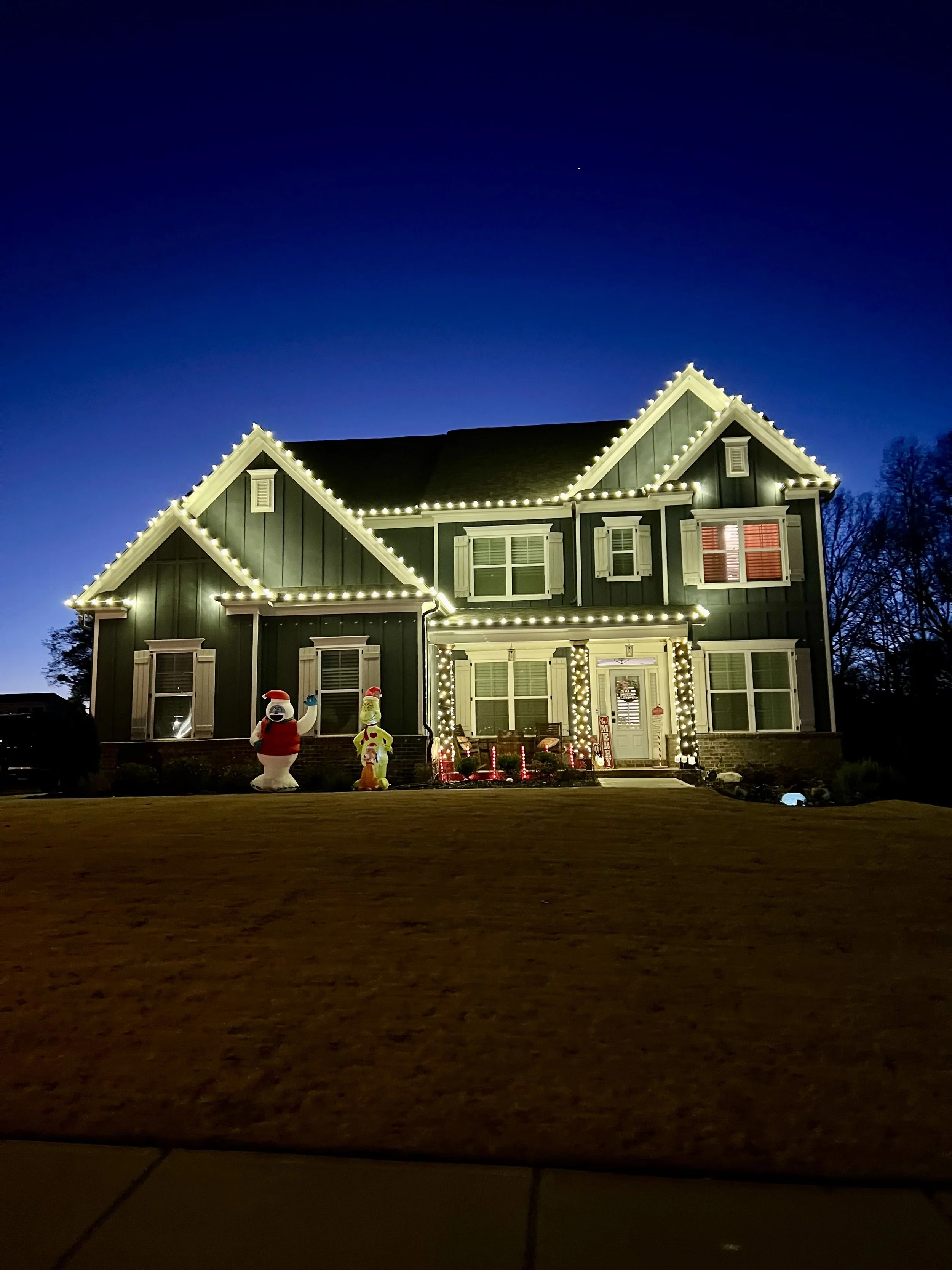 A two-story house decorated with white string lights along the roofline, windows, and porch. Inflatable Christmas decorations, including Santa Claus and a yellow character, are placed in the front yard. The house has white trim and green exterior. The sky is dark with a visible star.