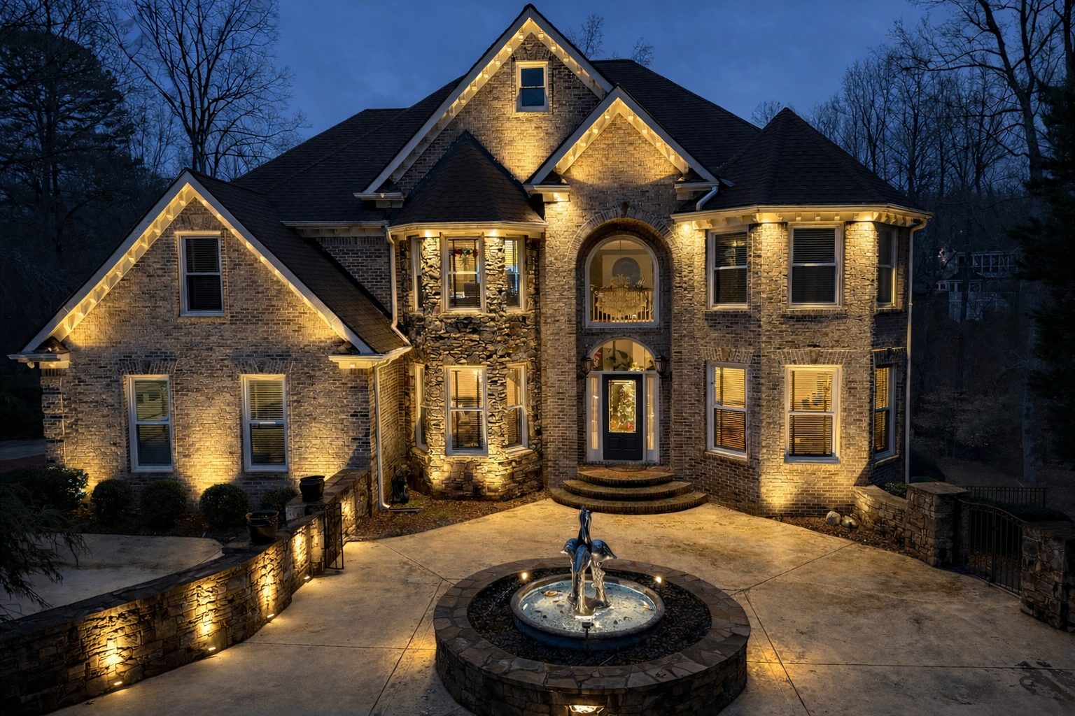 A large brick house illuminated at night with decorative outdoor lighting. Features multiple windows, a stone fountain with sculptures in the front yard, and a curved walkway with stairs leading to the front door. Trees are visible in the background.