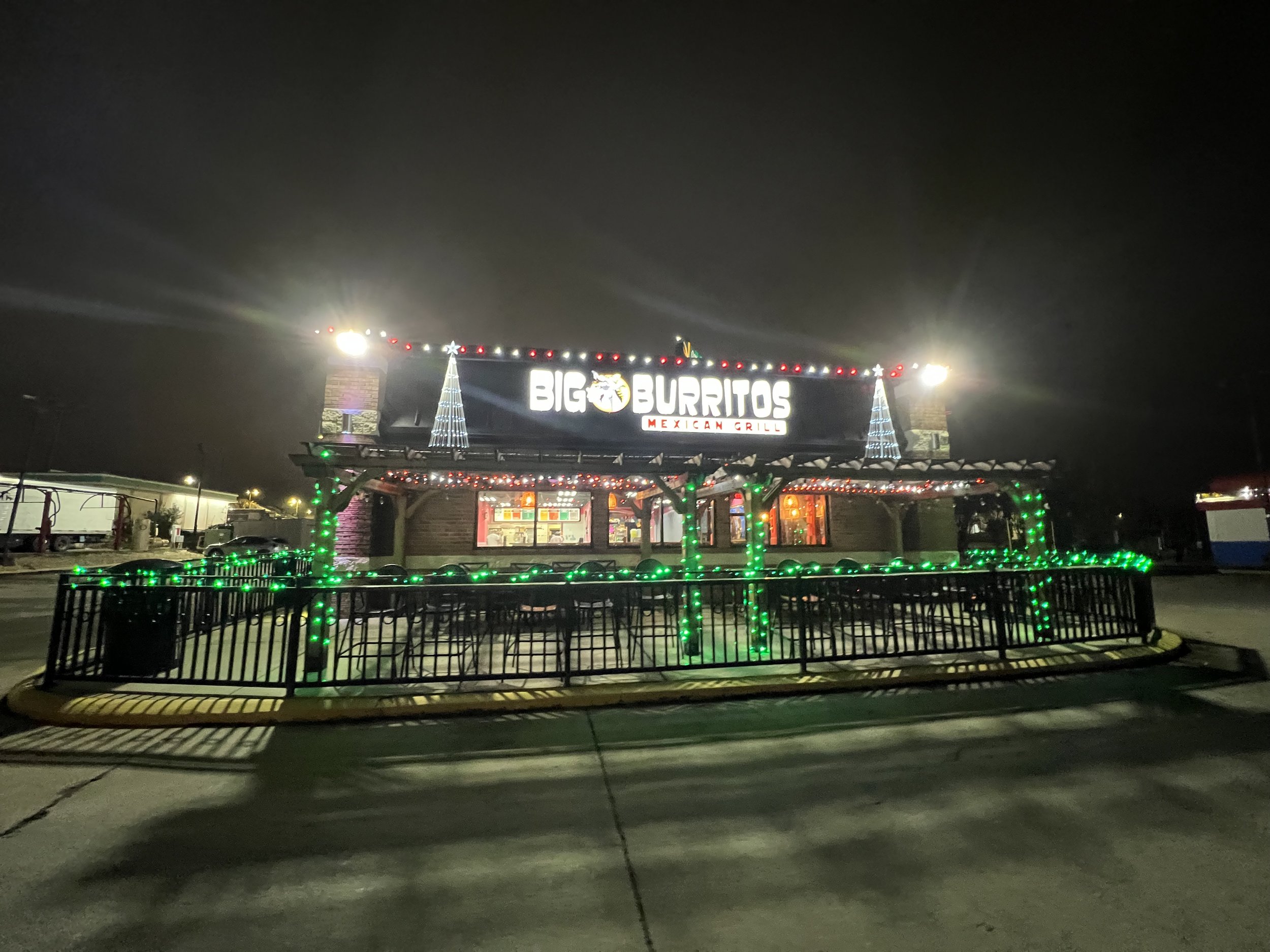 Nighttime scene of a Big Burritos Mexican Grill restaurant sign decorated with festive Christmas lights, visible from the parking lot with some cars and a fenced area in the foreground.