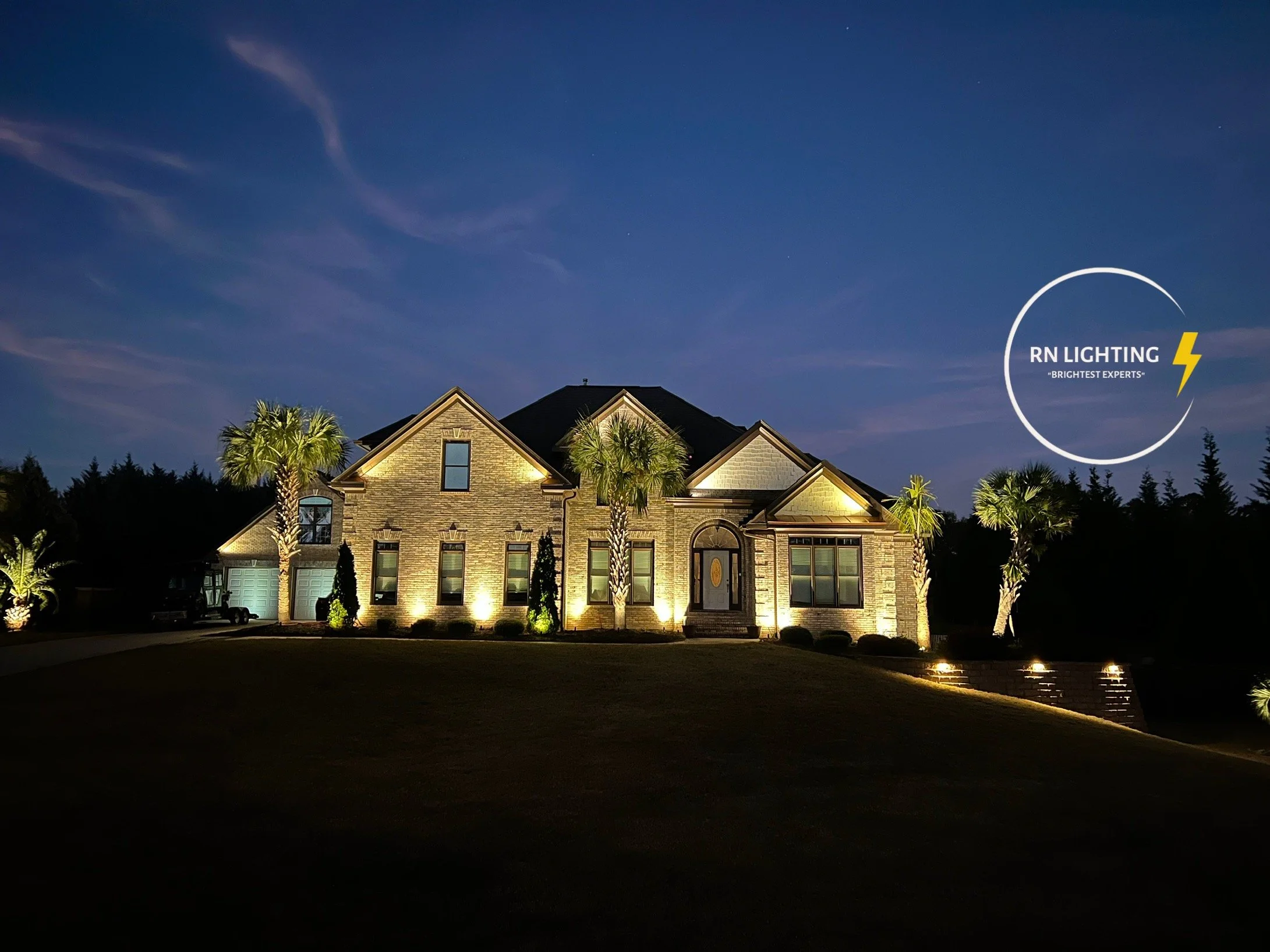 A large residential house illuminated with outdoor lighting at night. The house has brick walls, multiple gables, and a front door with windows. There are palm trees and a well-maintained lawn in the front yard, with some landscape lighting. The sky is clear with visible stars, and there is a logo of RN Lighting in the lower right corner.