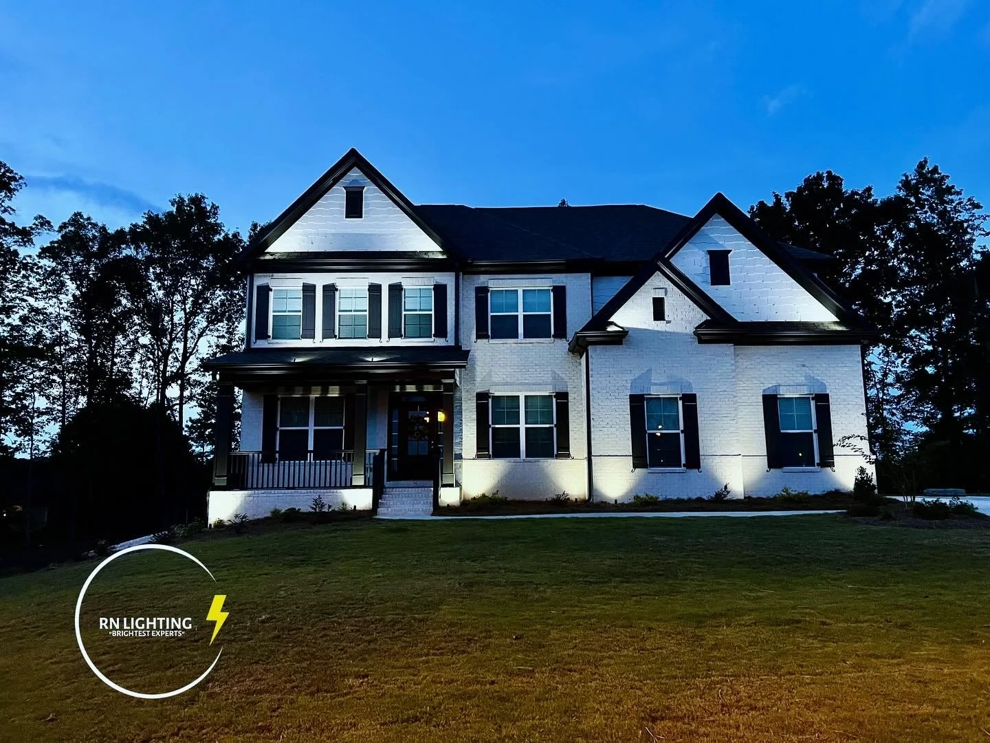 A large white two-story house with black window shutters and a front porch illuminated at night. Surrounding trees and a grassy lawn are visible, with the house demonstrating exterior lighting from RN Lighting.