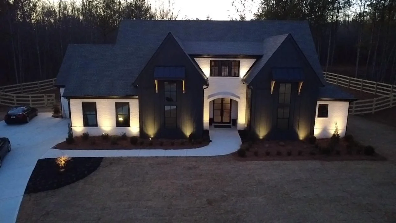 A modern two-story house at dusk with exterior lights. It has dark and white walls, multiple gabled roofs, large windows, a paved pathway leading to the front entrance, and a spacious driveway with parked cars. There is a wooden fence surrounding the property and trees in the background.