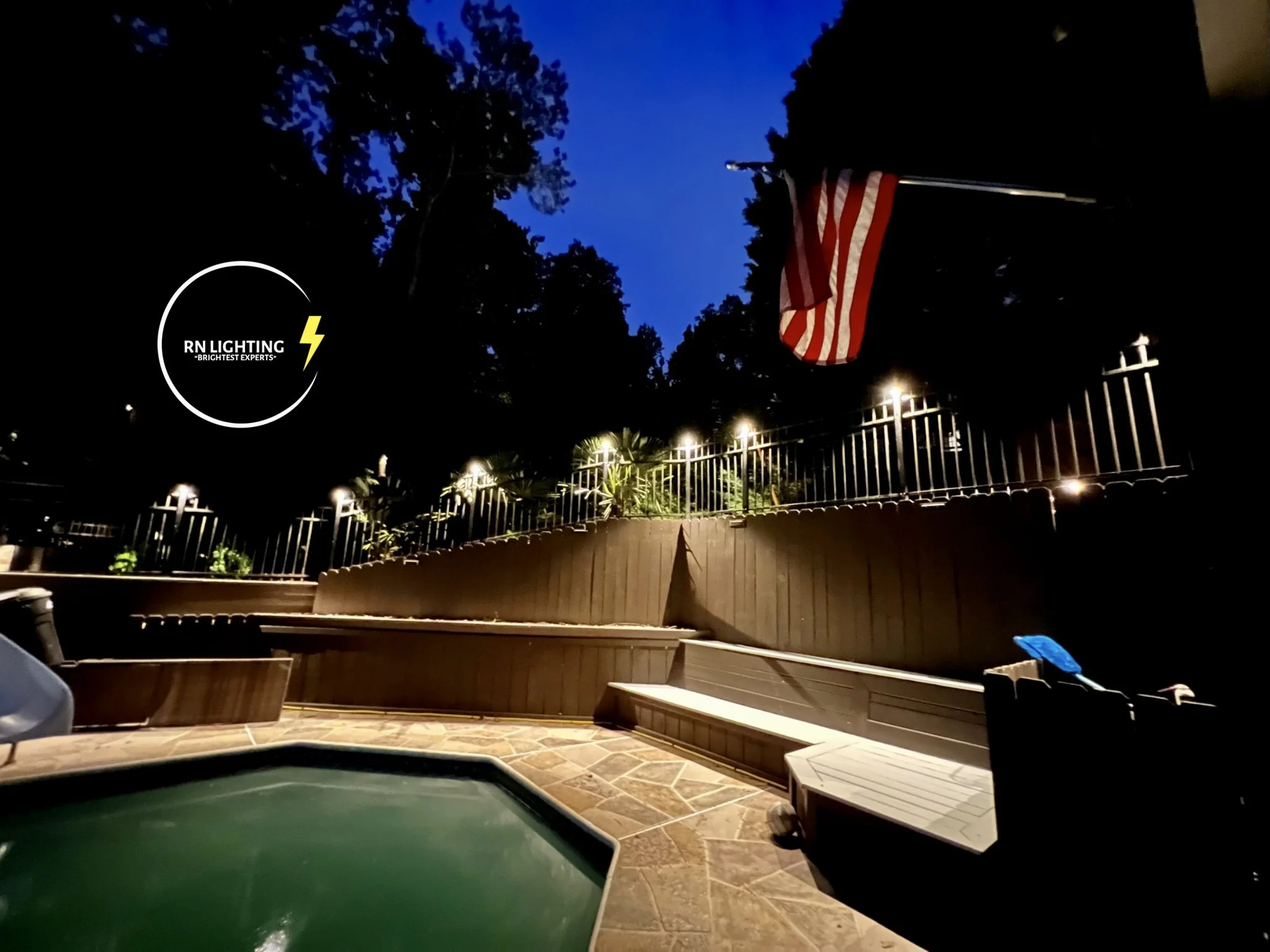 Backyard patio scene at dusk with a pool table in the foreground, outdoor lighting on a fence, an American flag hanging, and a cloudy evening sky.