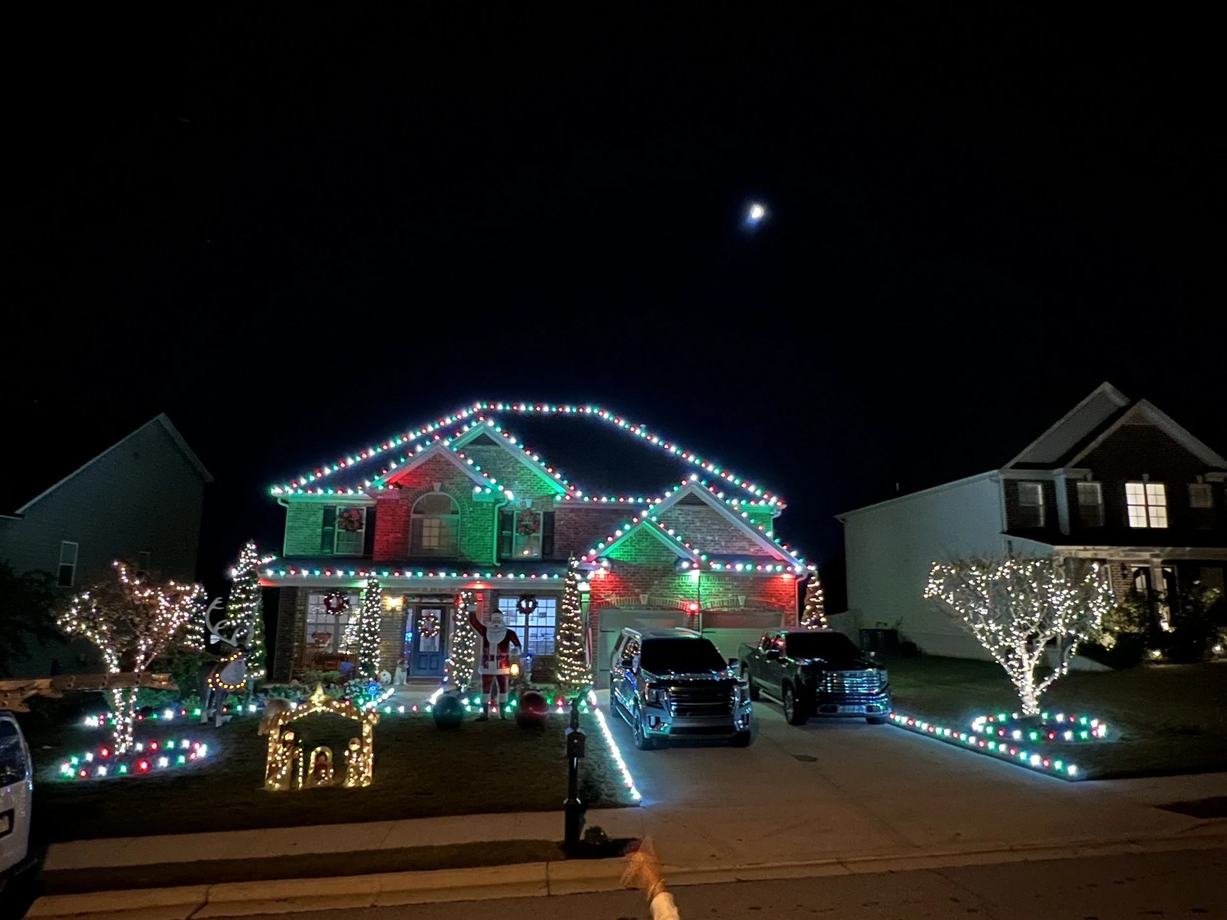 A house decorated with colorful Christmas lights, garlands, and festive displays at night, with a large illuminated moon in the sky.