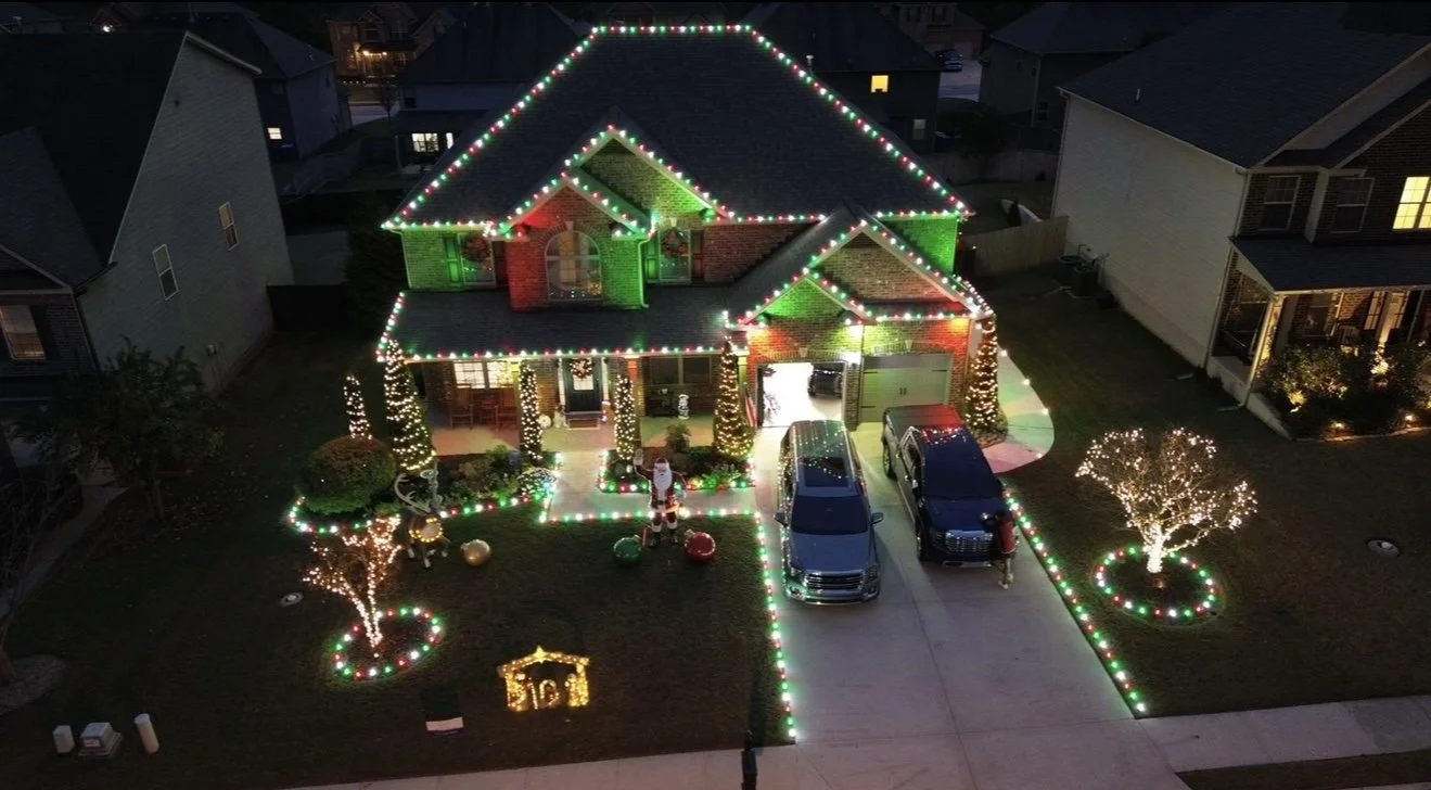 A house decorated with Christmas lights, with multicolored lights outlining the roof and the edge of the house. The front yard has lit-up trees, a nativity scene, and Christmas ornaments, creating a festive holiday display. There are three cars parked in the driveway.