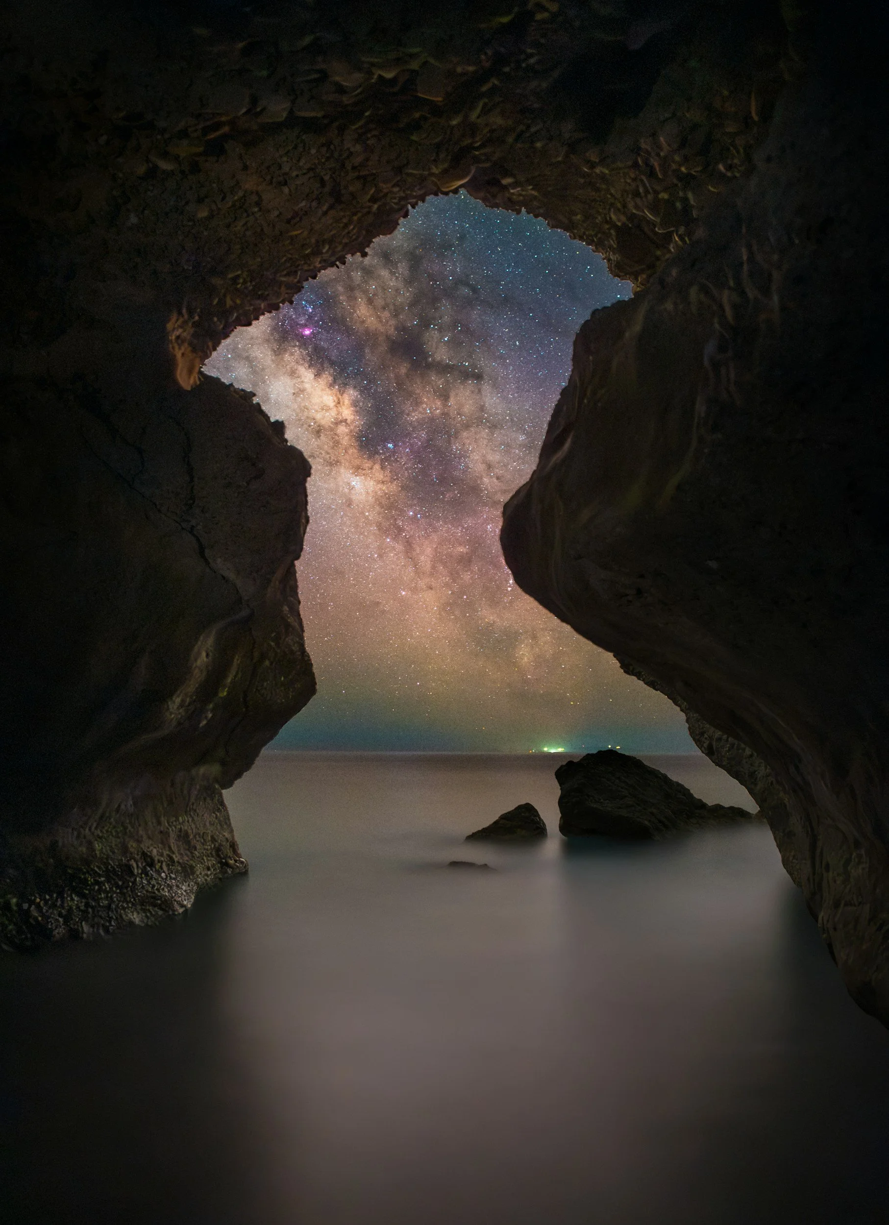 Nighttime view of the Milky Way galaxy through a rocky coast cave opening, with calm water and distant lights on the horizon.