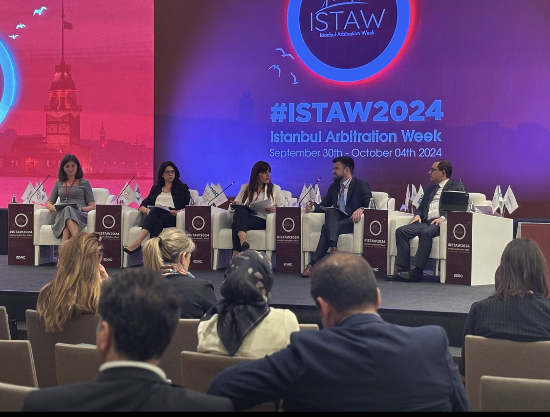 Panel discussion at Istanbul Arbitration Week 2024 with five speakers seated on stage in front of a large screen displaying event info, audience seated facing the stage.