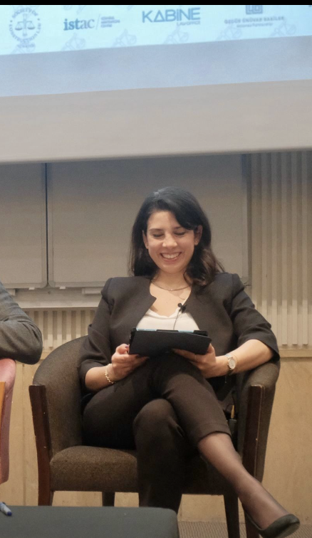 Smiling woman sitting cross-legged in a brown armchair, holding a tablet during a panel discussion or presentation.