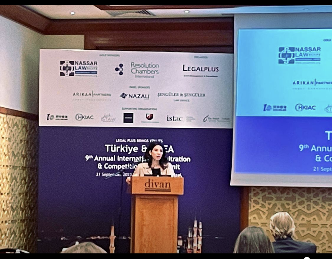 A woman stands at a wooden podium labeled 'divan' delivering a speech at a conference. Behind her is a blue backdrop with various sponsor logos and the event title, which includes 'Türkiye & USA' and mentions the 9th Annual International Arbitration & Competition Summit. Attendees are seated in front of her, and a large screen to her right displays event details.