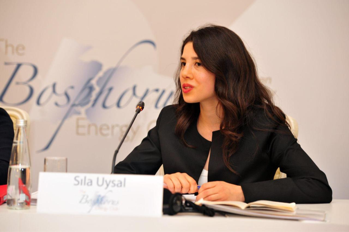 A woman with long dark hair, wearing a black blazer, is speaking into a microphone at a conference table. There is a nameplate in front of her reading "Sila Uysal" and a notebook, along with a glass of water and a bottle on the table.