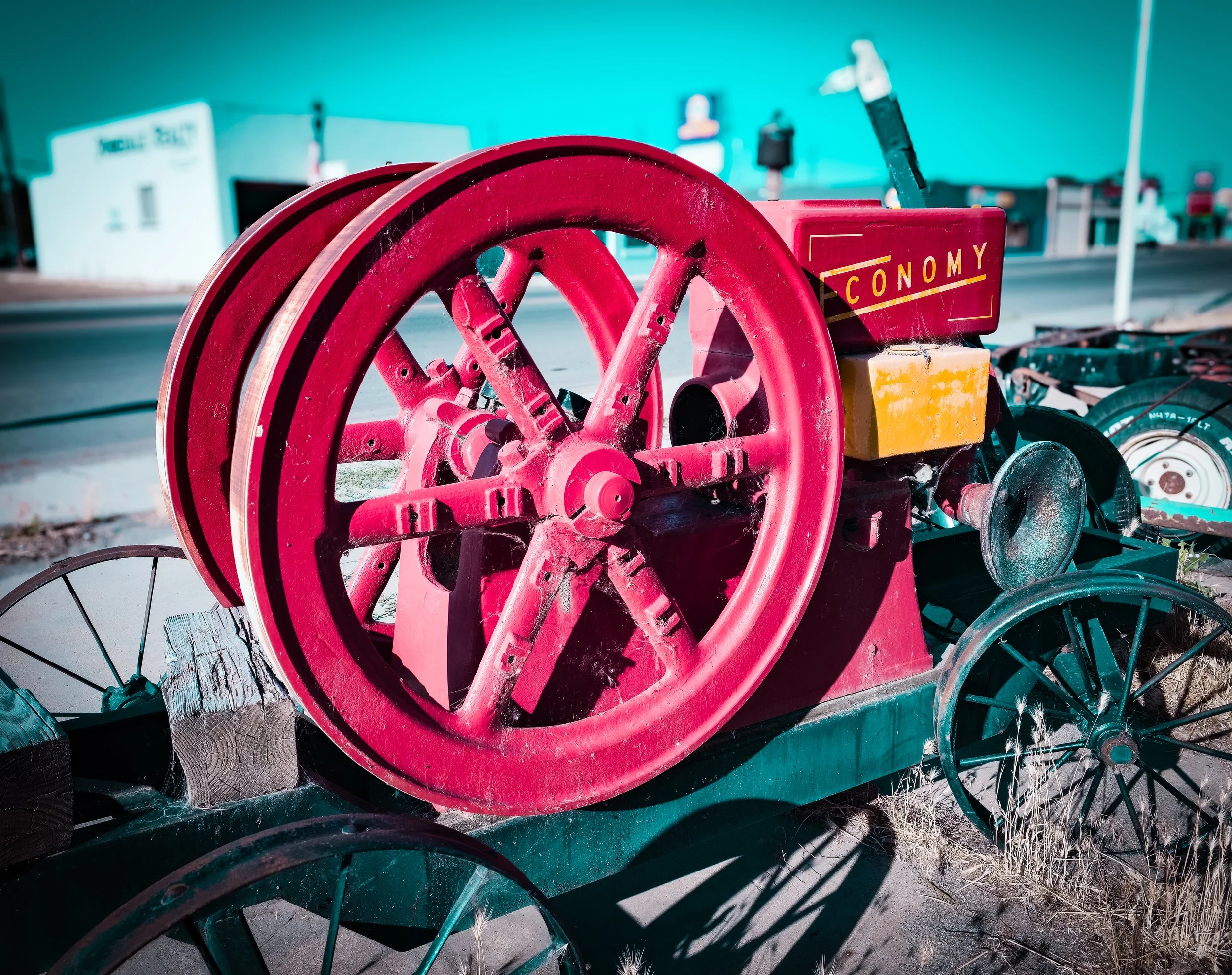 Close-up of a red vintage bicycle wheel and part of a yellow box on a wooden cart in an outdoor area with a blurred teal wall and signage in the background.