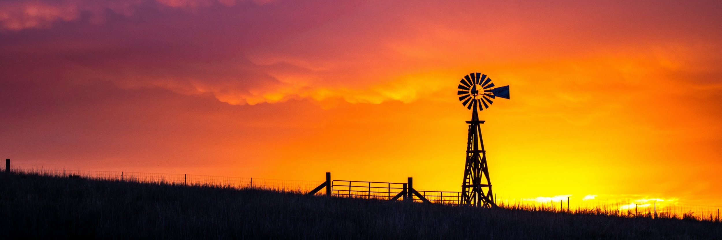 Silhouette of a windmill against a vibrant orange and pink sunset sky, with a grassy foreground and a fence.