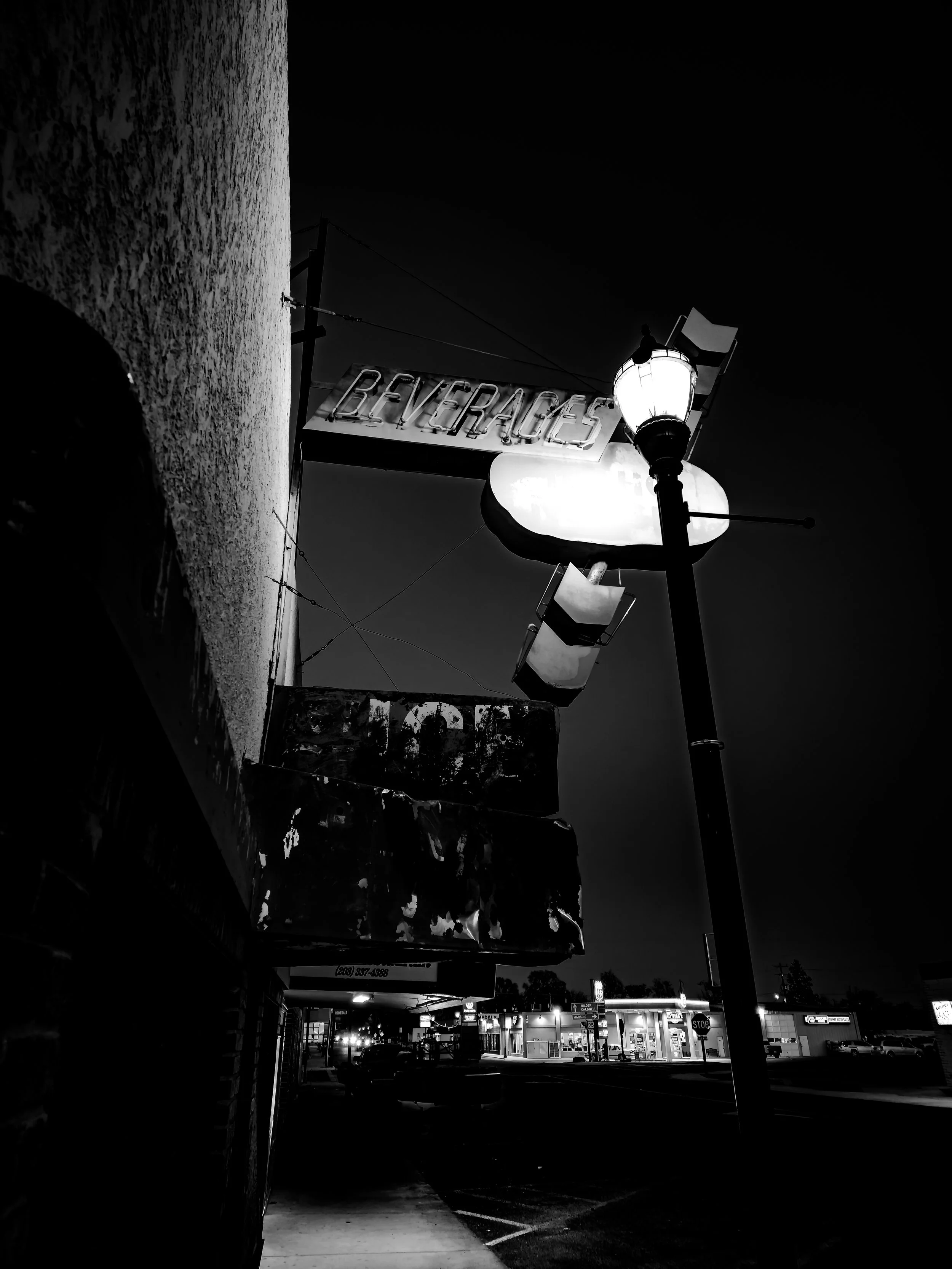 Nighttime street view with illuminated sign reading 'Beverages' and a vintage street lamp, storefronts, cars, and a sidewalk in the background.