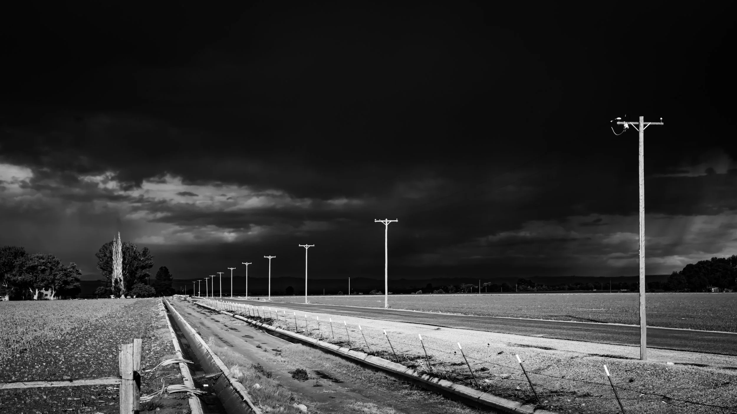 Black and white photo of an empty rural road with utility poles stretching into the distance, dark stormy clouds overhead, and trees on the left side.