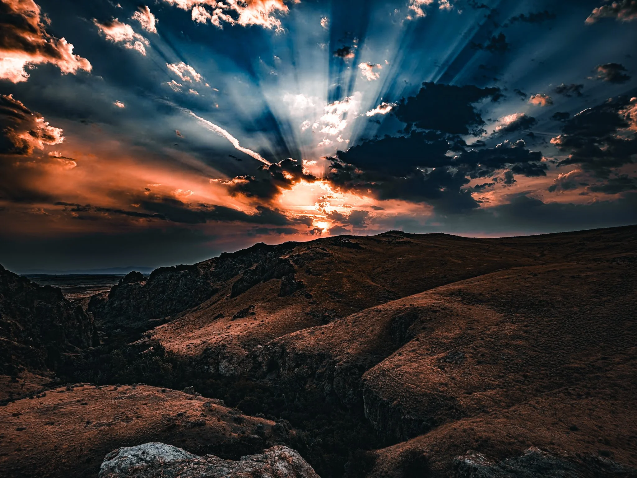 Sunset over rugged hills with clouds and sun rays breaking through, creating a dramatic sky.