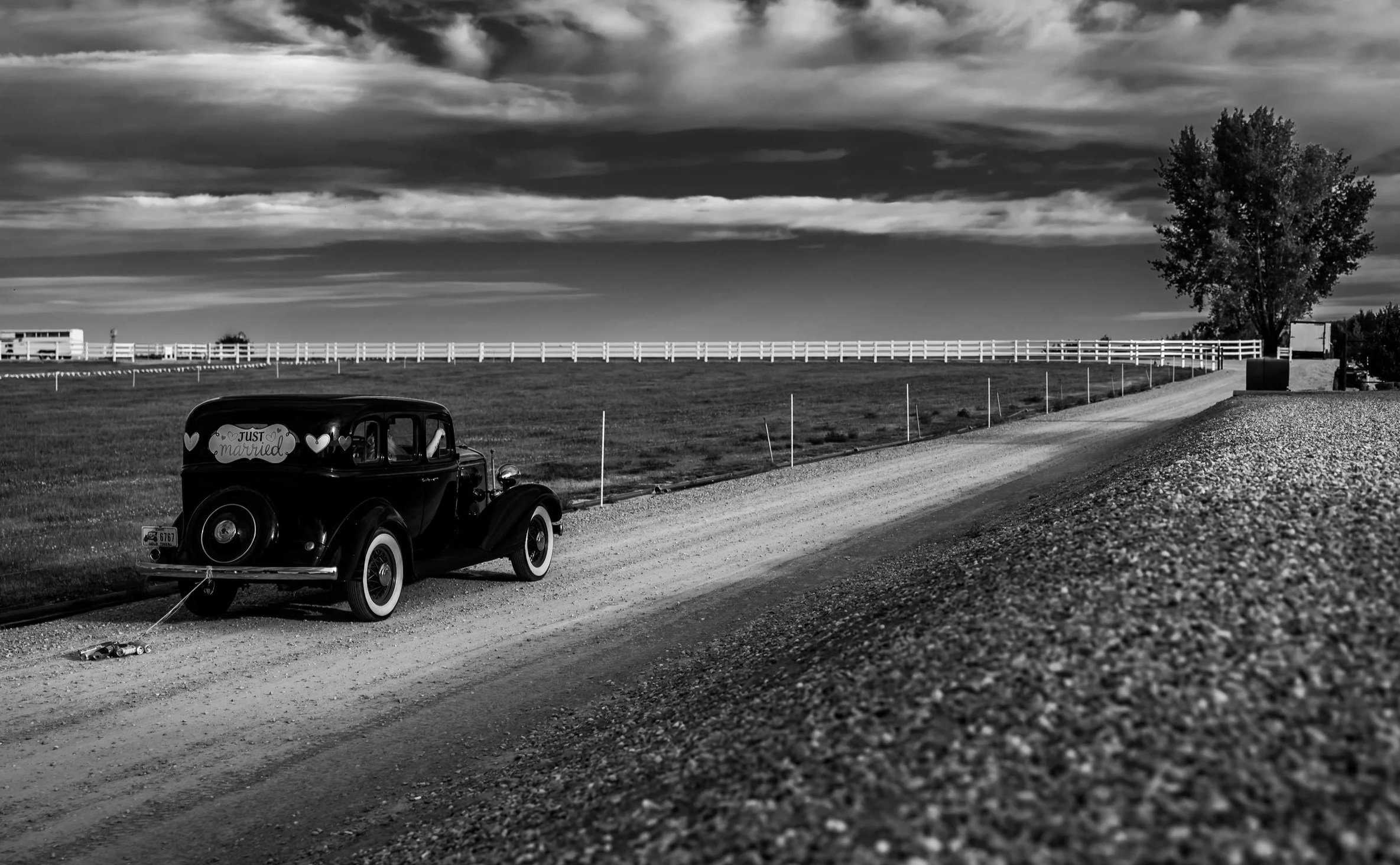 A vintage car with a 'Just Married' sign in the window parked on a dirt road in a rural area, with a large tree, a white fence, and a cloudy sky in the background.