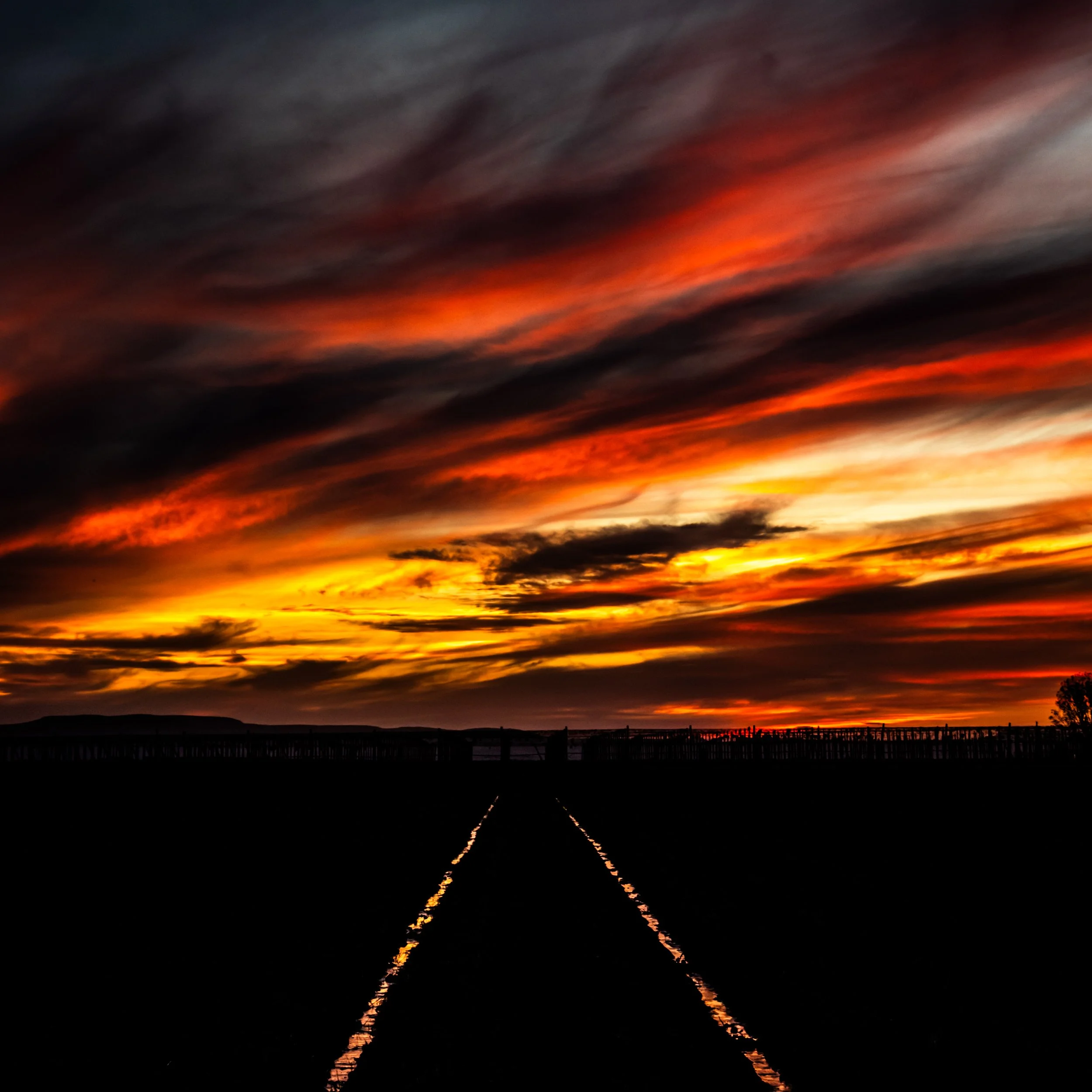 A sunset sky with dark clouds and warm orange, red, and yellow hues, with silhouettes of trees and a railroad track in the foreground.