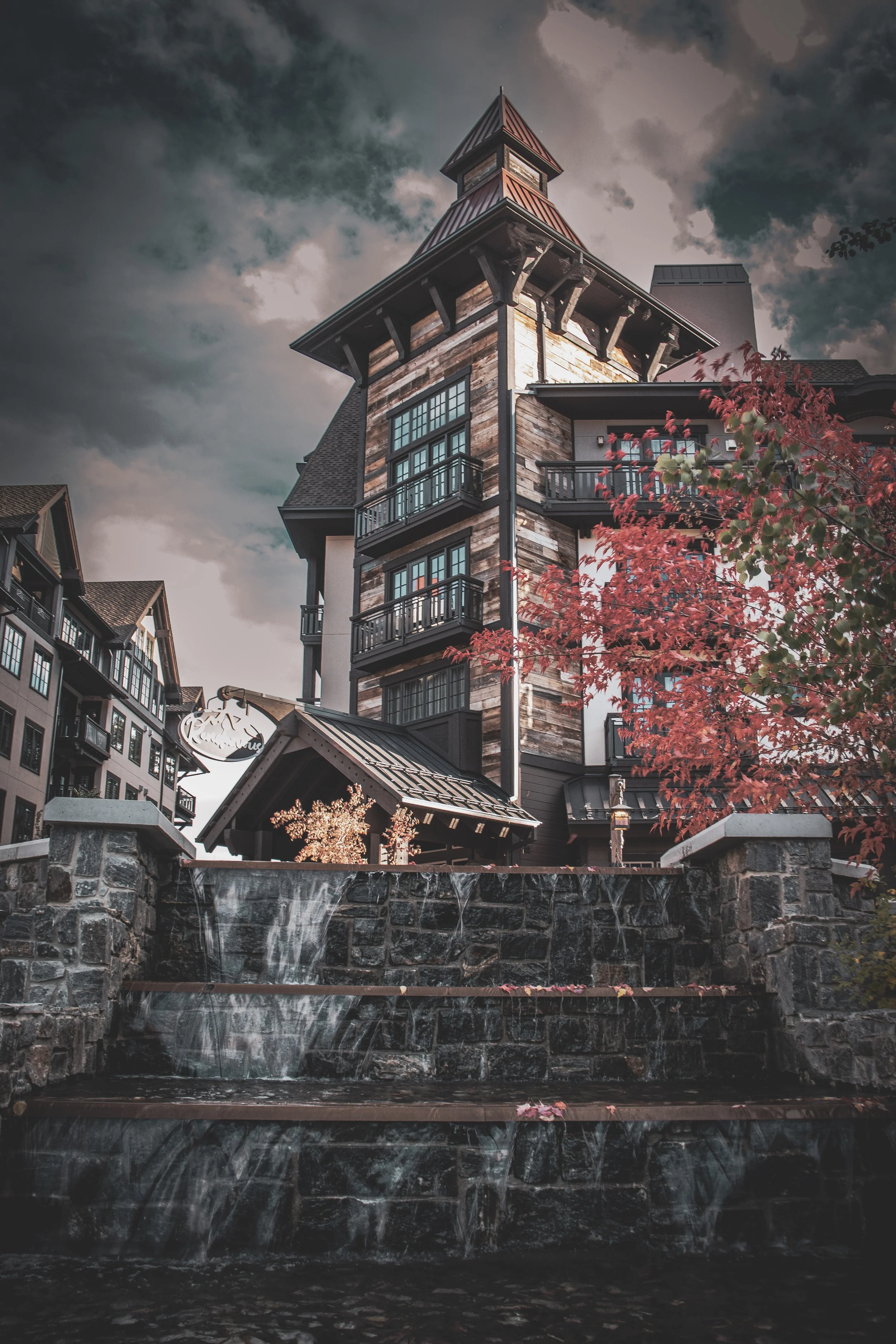 A multi-story building with a steep roof and a tower, featuring large windows and balconies, situated near a water feature with waterfall and stones, and surrounded by trees with red and green leaves under a cloudy sky.