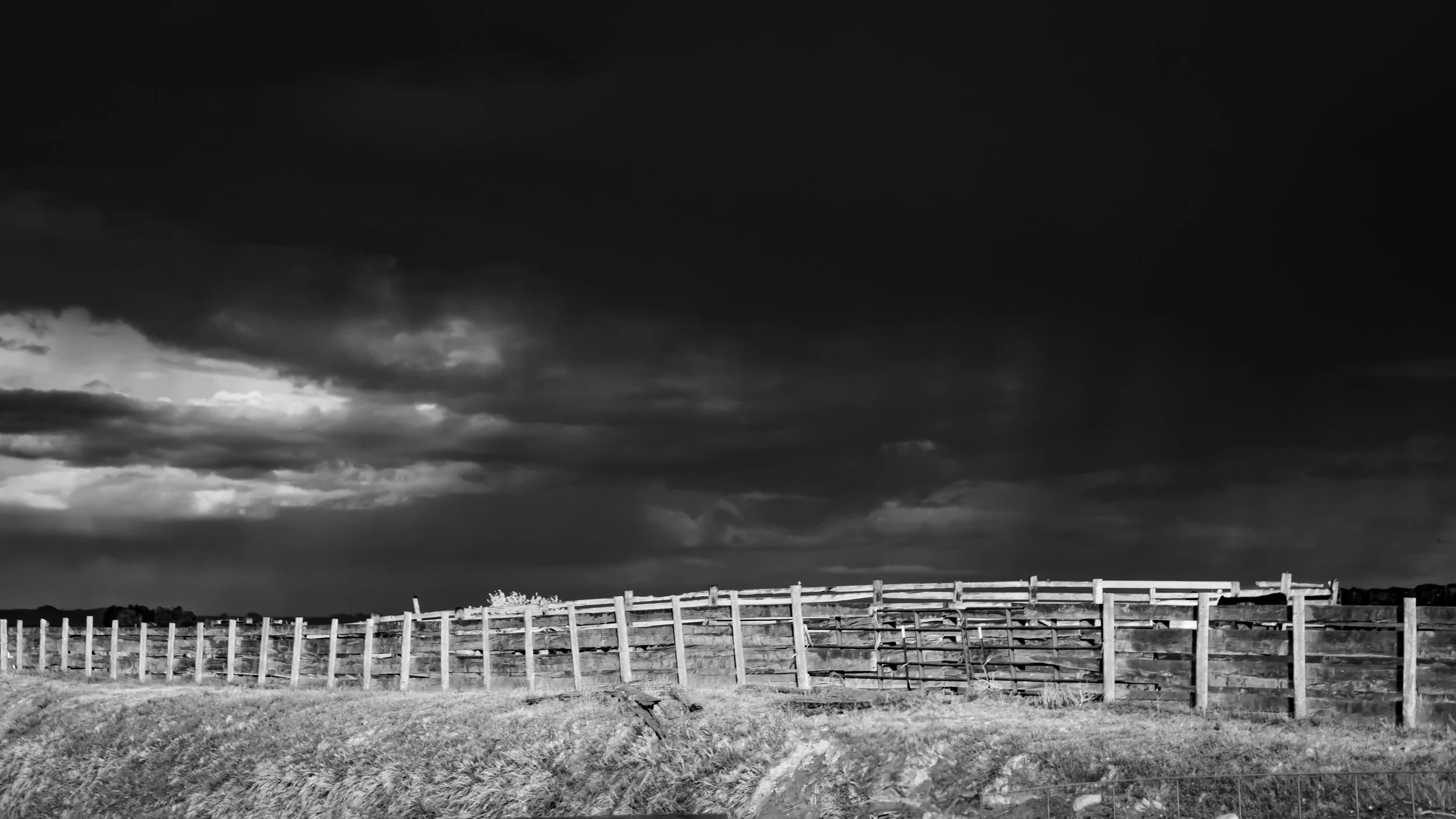 Black and white photo of a farm field with a wooden fence and dark stormy clouds in the sky.