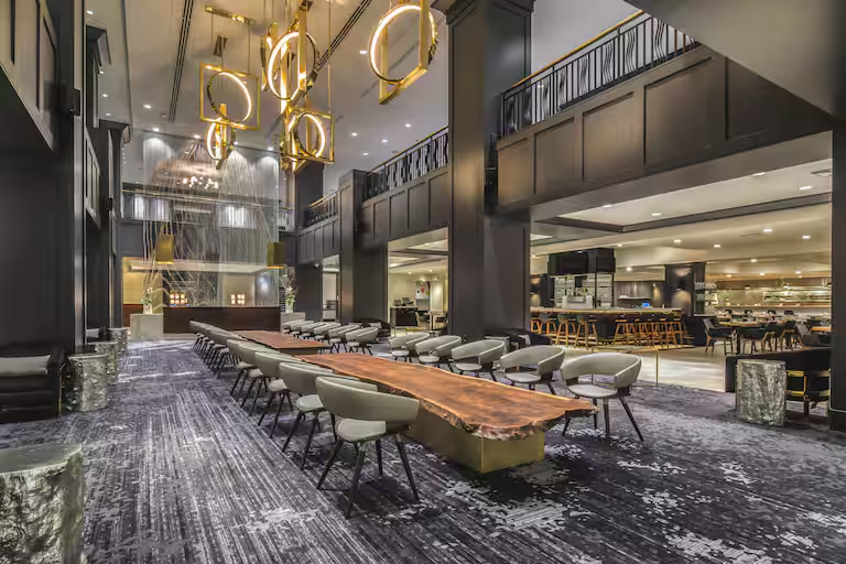 Elegant hotel lobby with high ceilings, modern chandeliers, a large wooden table surrounded by chairs, and a bar area in the background.