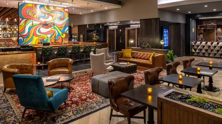 A stylish lounge area with various chairs and sofas, a patterned area rug, small lit candles, a bar with a colorful abstract painting, and a dark wood-paneled wall with an elevator.