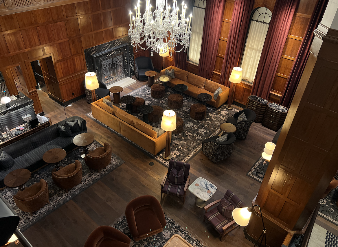 An elegant, spacious hotel lobby with wooden paneled walls, a large chandelier, various seating options including sofas and armchairs, and multiple lamps providing warm lighting.