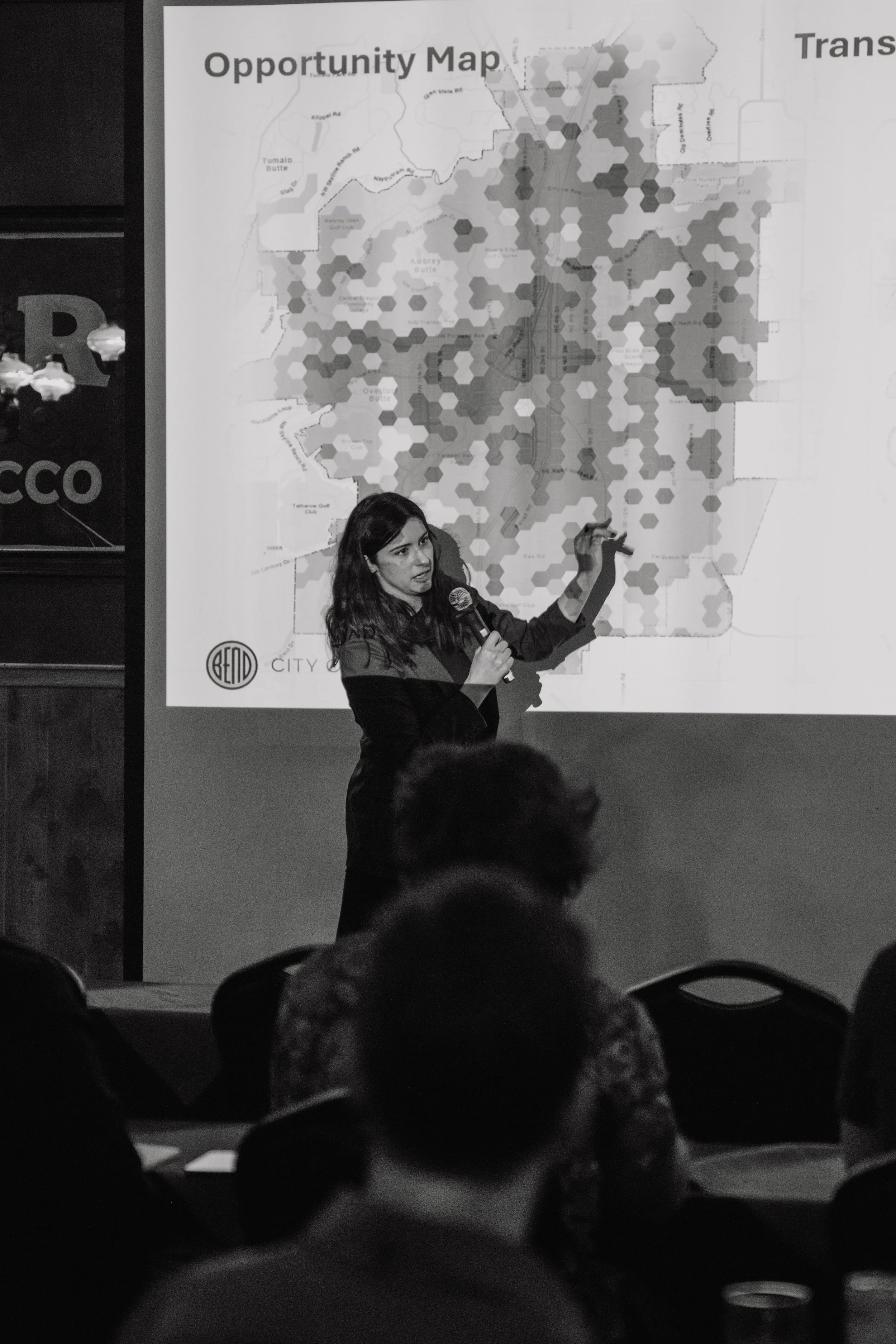 A woman is giving a presentation in front of a large map labeled 'Opportunity Map' with various shaded hexagons. She holds a microphone and points at the map with her right hand. Audience members are visible in the foreground.