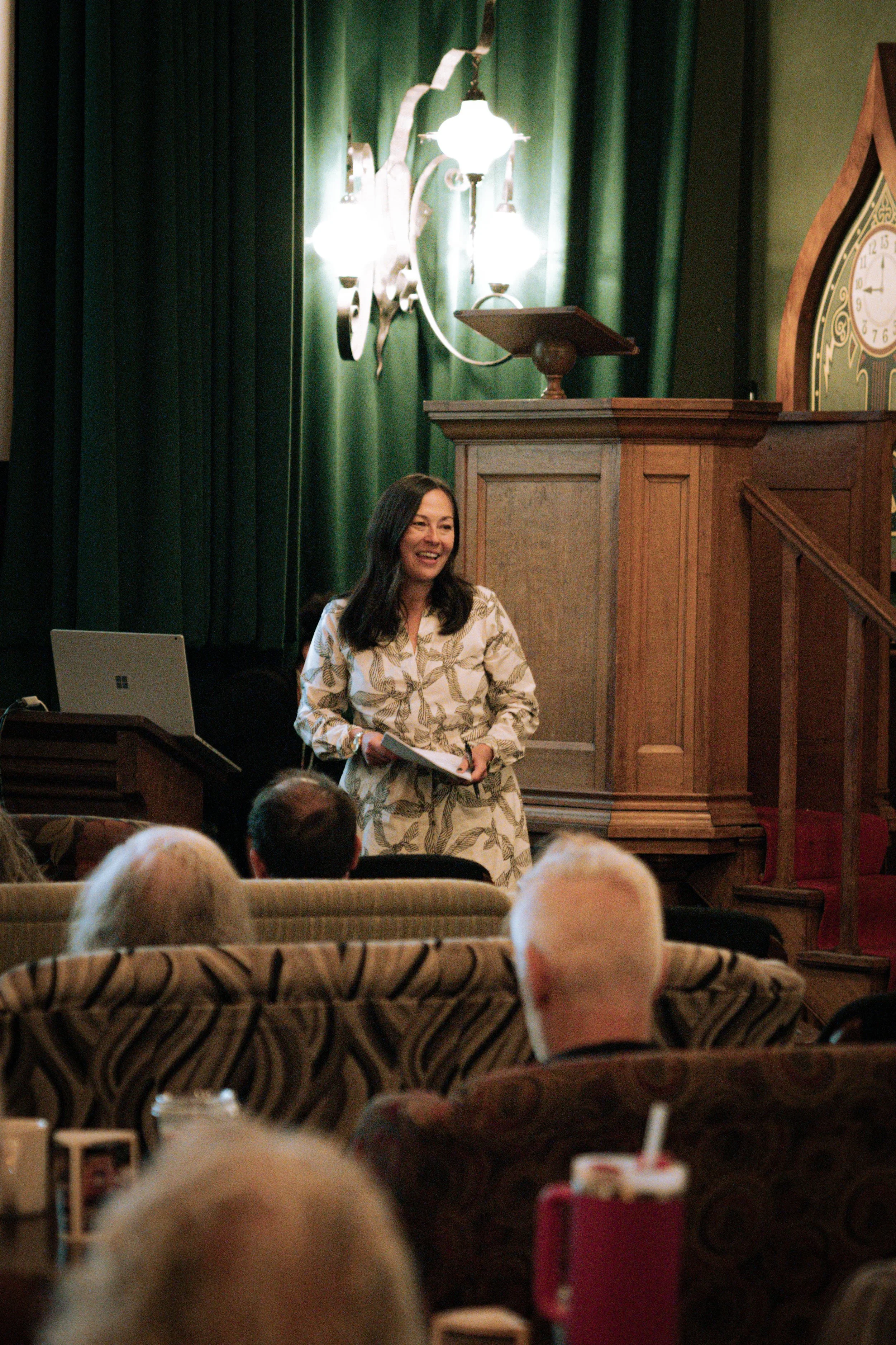 A woman with long dark hair, wearing a beige patterned dress, is standing and smiling while holding papers in a room with green curtains, wood-paneled wall, and a clock. There are audience members in the foreground, and a laptop and drinks on the table.