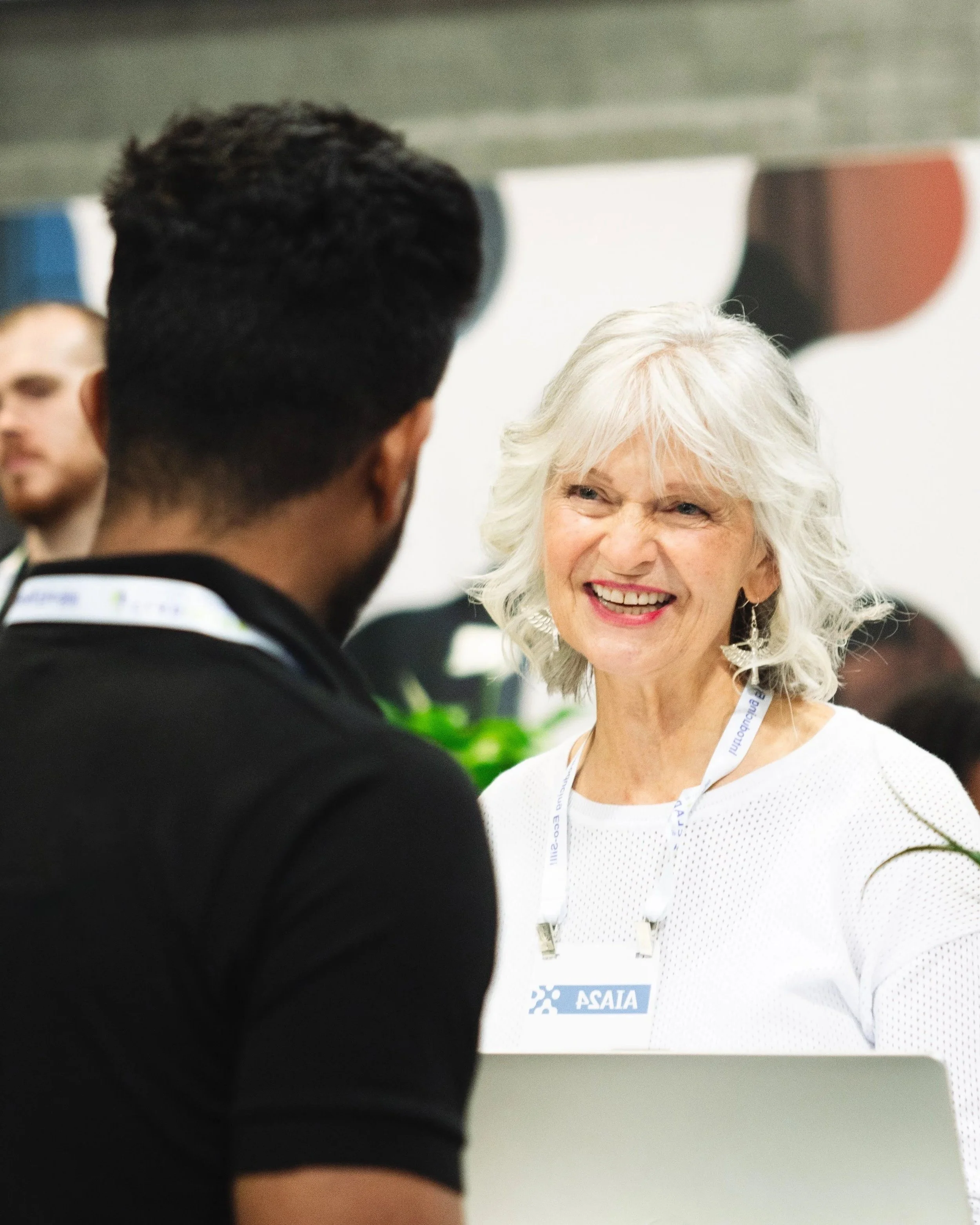 A woman with white hair and earrings smiling at a man with short dark hair at an indoor event, with a badge that says 'ASAIA' and a blurred background with other people.