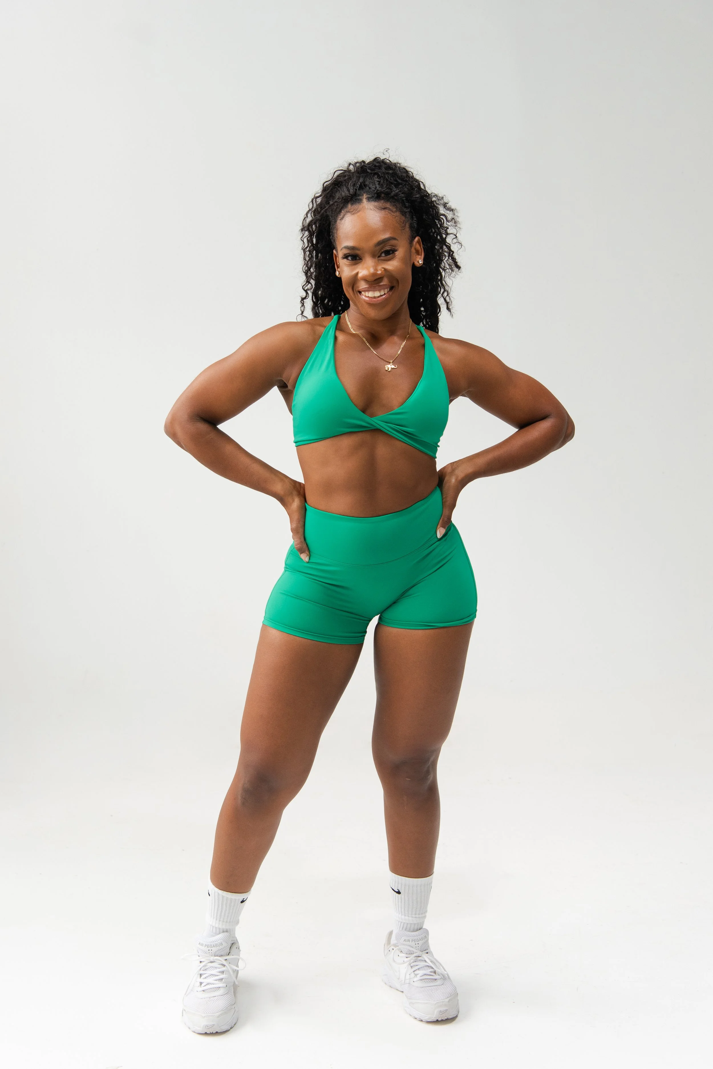 A young woman poses confidently, wearing a green athletic crop top and matching shorts, with white Nike sneakers and Nike socks, standing against a plain white background.