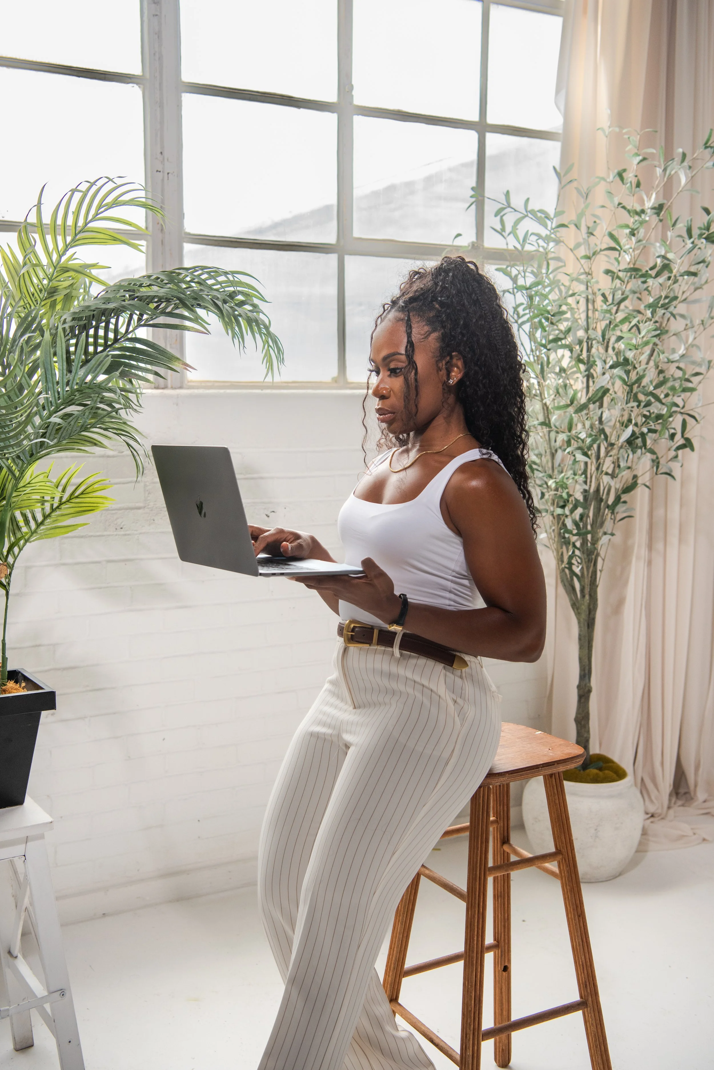 A woman with dark skin and curly hair tied in a ponytail stands indoors holding a laptop with both hands. She is dressed in a white tank top and high-waisted, white and beige striped pants. There are green plants around her, a large window with white framing behind her, and a wooden stool she is leaning on.