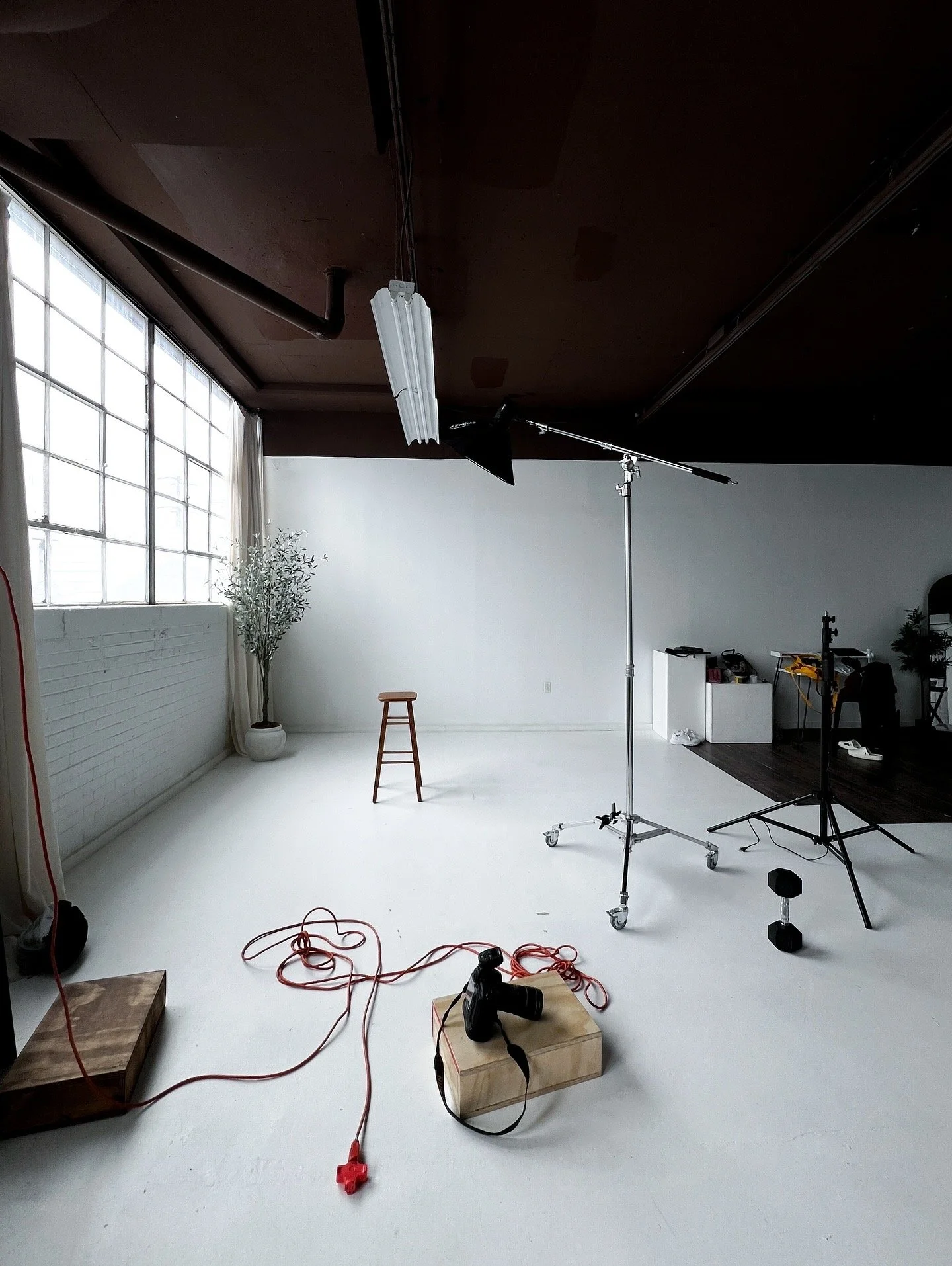 Photography studio with large white backdrop, studio light with reflector, camera on wooden platform, black weight, red extension cord, small stool, potted plant, and equipment on black table, window with grid glass panels in the background.