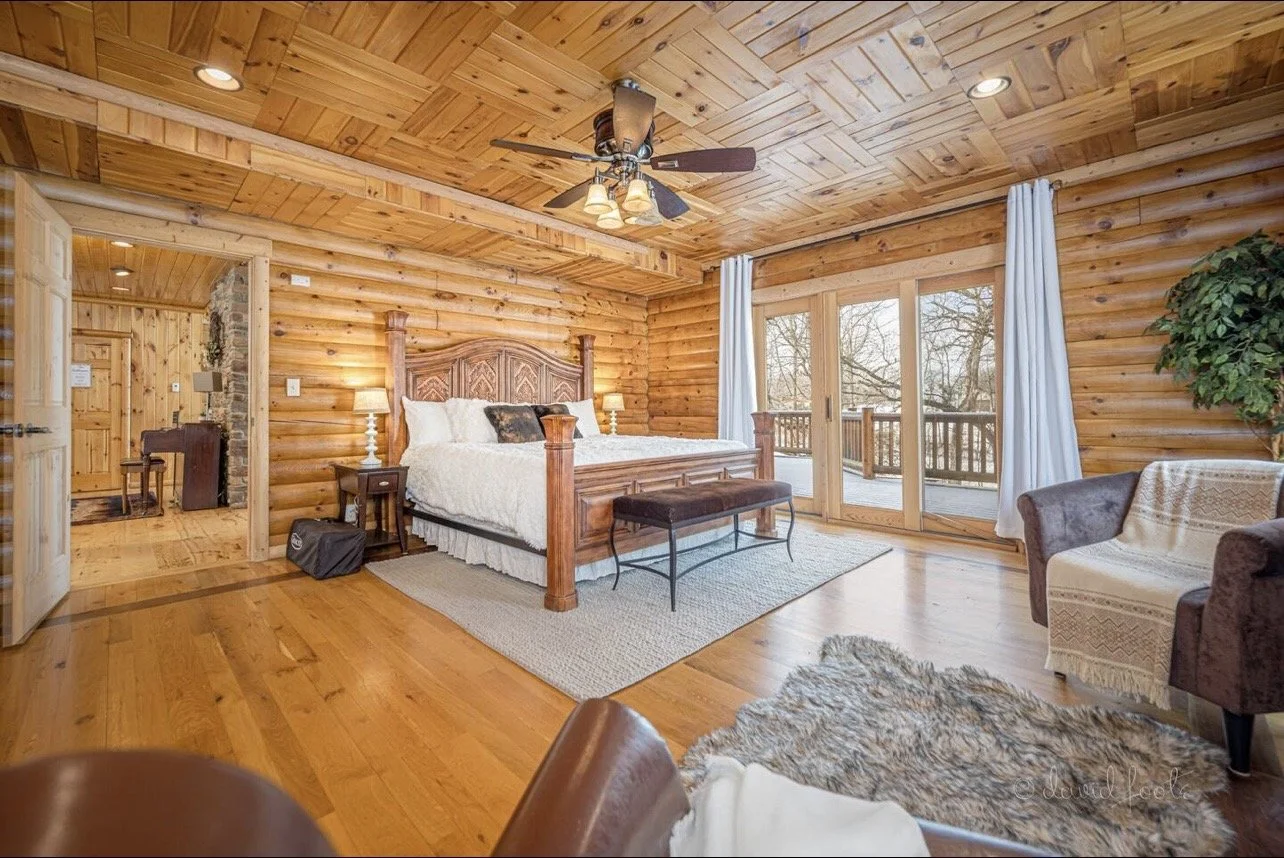 A cozy bedroom with wooden log walls, a large wooden bed with white bedding, a bench at the foot of the bed, two lamps on nightstands, a ceiling fan, a sliding glass door leading to a balcony, and a small sitting area with a gray armchair and a potted plant.