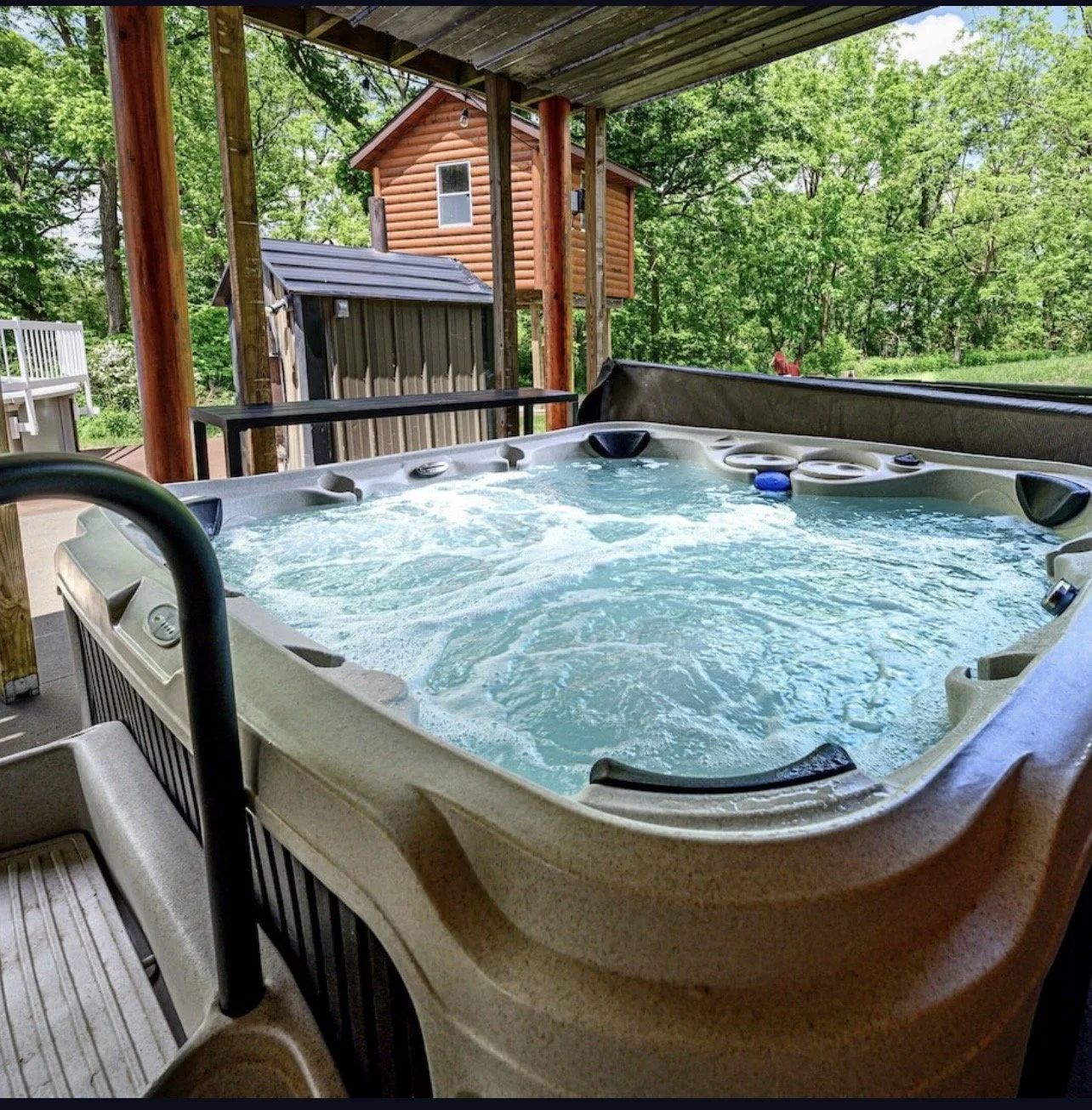 A hot tub filled with bubbling water on a covered deck, overlooking a backyard with green trees and a small wooden shed.