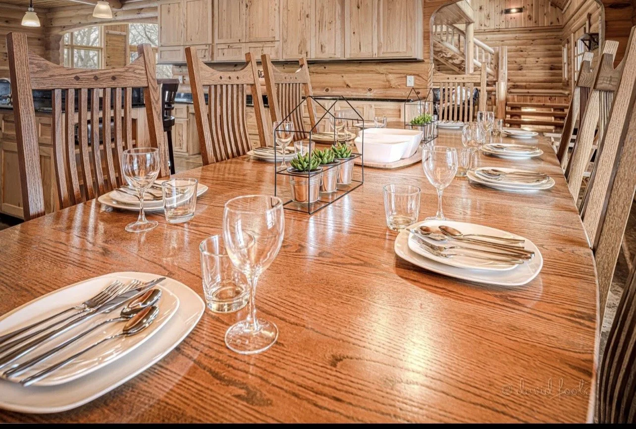 A large wooden dining table set for a meal, with plates, silverware, wine glasses, water glasses, and a decorative centerpiece with small potted succulents inside black metal frames, in a wood-paneled room with natural light and stairs in the background.