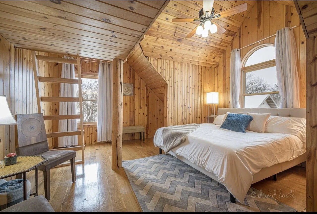 A cozy bedroom with wooden walls and ceiling, a large bed with white bedding and pillows, a ceiling fan with lights, a window with white curtains, a small bench, and chairs. The room has a warm, rustic feel.