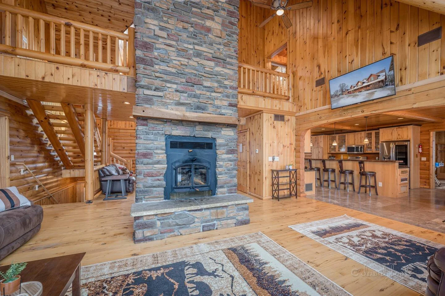 Interior of a rustic wooden living room with a stone fireplace, wood-paneled walls, a flat-screen TV, and a kitchen with a bar counter and chairs.