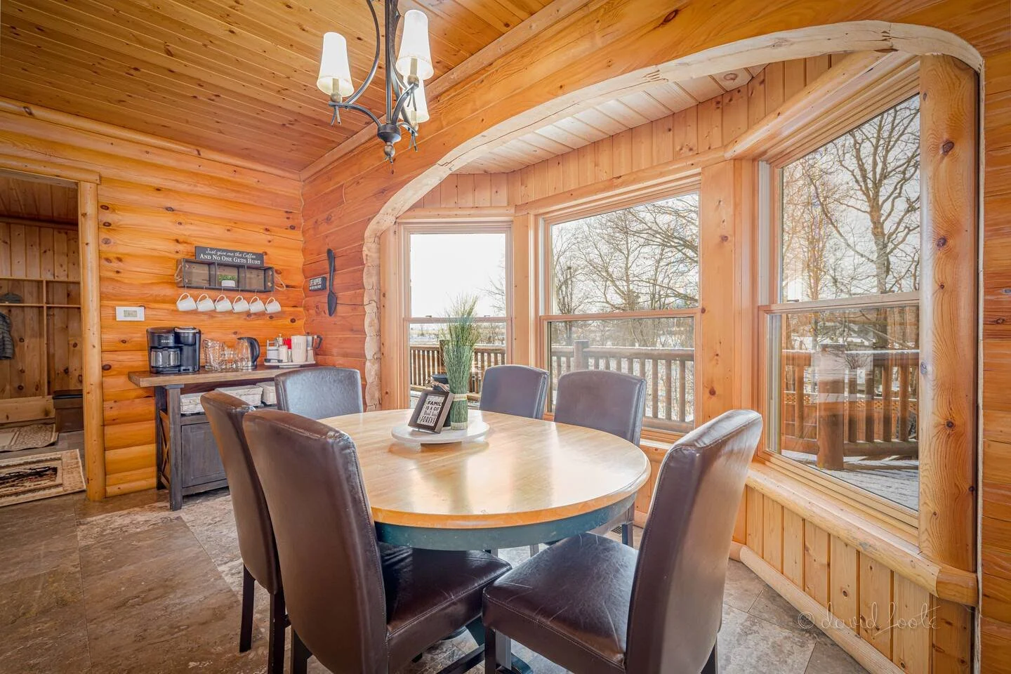 Dining area with wood-paneled walls and large windows, featuring a round wooden table with six leather chairs, a chandelier overhead, and a small side table with coffee mugs and a coffee maker.
